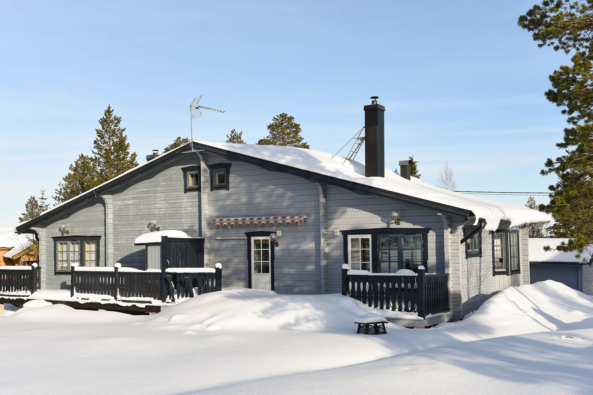 En hyggelig grå hytte i Wagrain, med snedækket tag og veranda, omgivet af træer under en klar blå himmel. Hytten har en skorsten og en antenne med sne, der hober sig op på jorden og udendørsmøbler - perfekt til et skiferie-tilflugtssted.