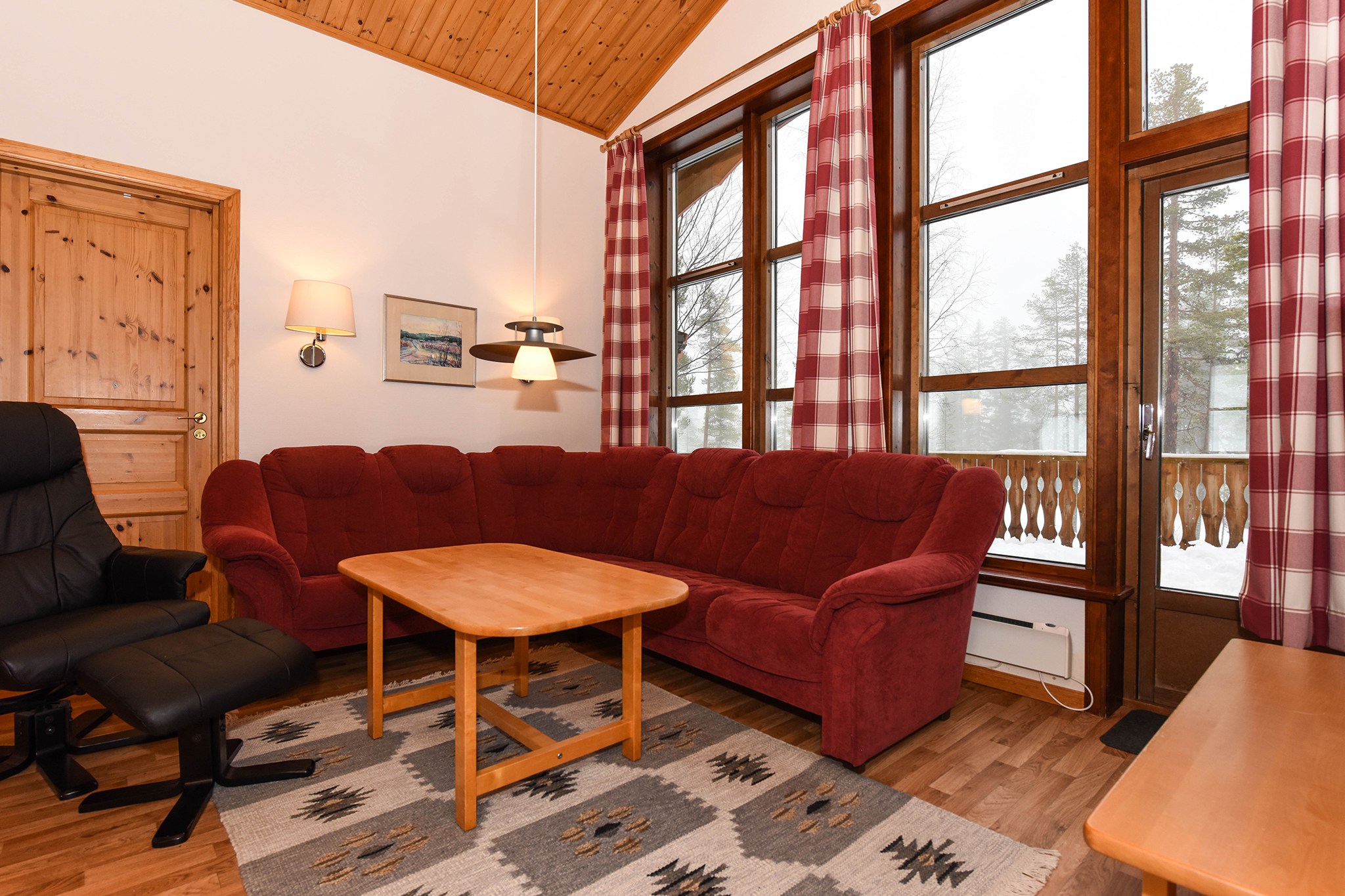 En hyggelig stue med træloft og trægulv minder om Saalbach, med en rød hjørnesofa, en sort lænestol, et sofabord i træ og et mønstret tæppe. Store vinduer med rødternede gardiner giver udsigt til snefyldte træer udenfor. Perfekt til dit skiferie-retreat!.
