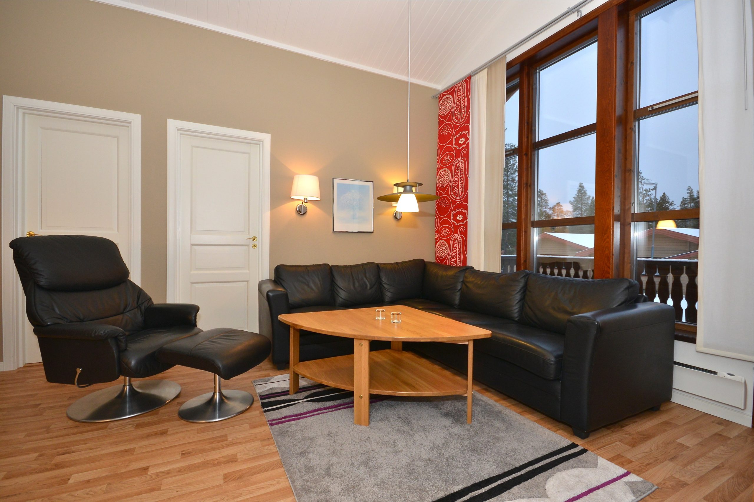En hyggelig stue, der minder om et Saalbach-retræte, har en stor sort lædersofa, en lænestol med fodstøtte og et sofabord i træ på et mønstret tæppe. Lysebrune vægge prydet med to væghængte lamper lyser varmt, mens høje vinduer med røde gardiner slipper naturligt lys ind.
