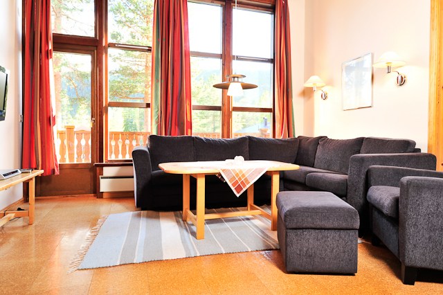 En hyggelig stue, der minder om et Wagrain-retræte, har en mørkegrå sektionssofa med et sofabord i træ og en skammel. Store vinduer med røde gardiner lukker naturligt lys ind, og der er en lille altan udenfor. Et stribet tæppe tilføjer et strejf af farve til det indbydende rum.