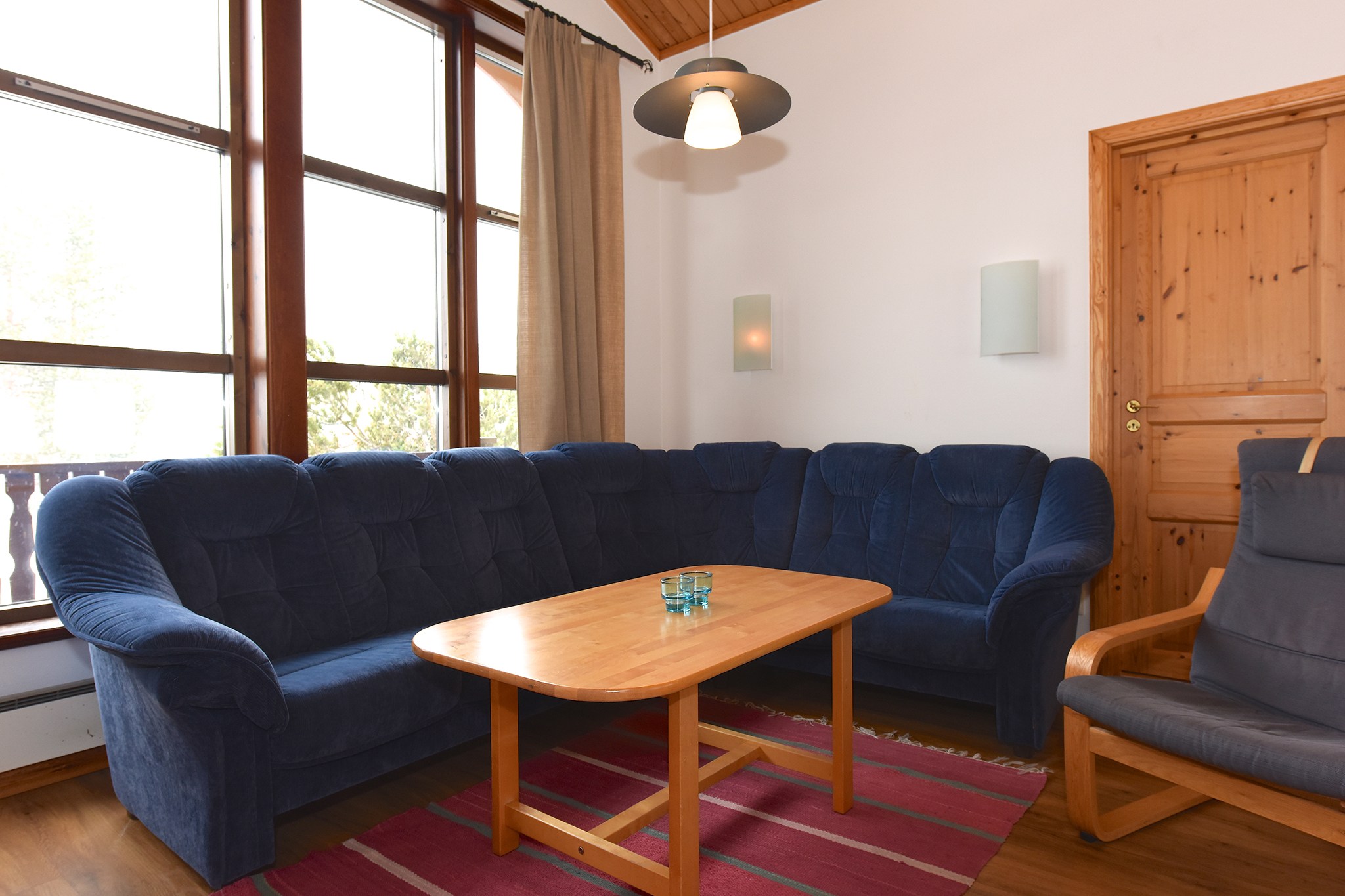 En hyggelig stue med en stor blå hjørnesofa omkring et sofabord i træ fremkalder charmen ved Westendorf. En trælænestol og rødt tæppe giver varme, der minder om et sommerferie Østrig-retræte. Høje vinduer indbyder til naturligt lys, mens et hængende armatur oplyser rummet ved siden af trædøren til højre.