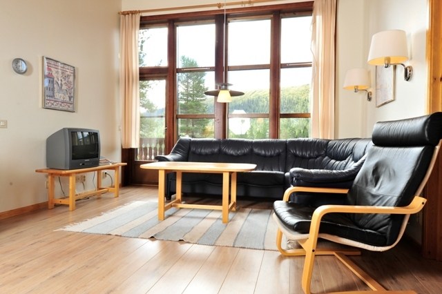 En hyggelig stue med et stort vindue indbyder naturligt lys, der minder om en Westendorf skiferie. Den har en sort lædersofa og lænestol, et sofabord i træ, væglamper og et dekorativt tæppe på trægulvet. Uden for vinduet fuldender en naturskøn udsigt den rolige stemning..