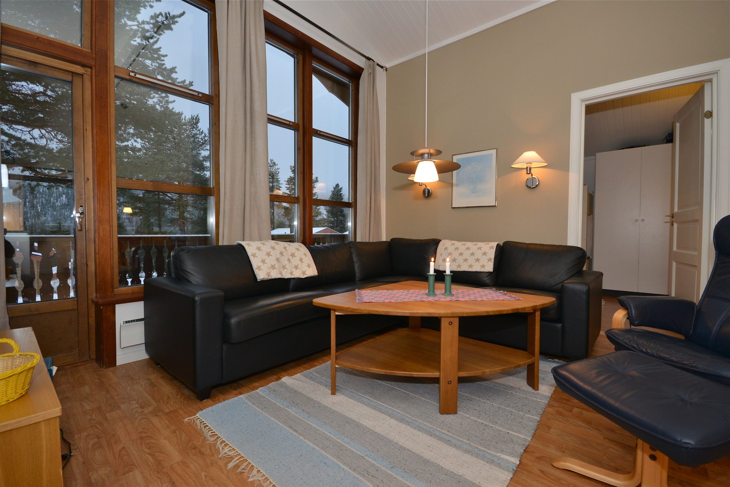 Hyggelig stue med sort hjørnesofa i læder, rundt sofabord i træ og to tændte stearinlys. Store vinduer afslører et snelandskab, der minder om Saalbach udenfor. En flettet kurv sidder ved siden, og en lille væglampe tilføjer varme til rummet.