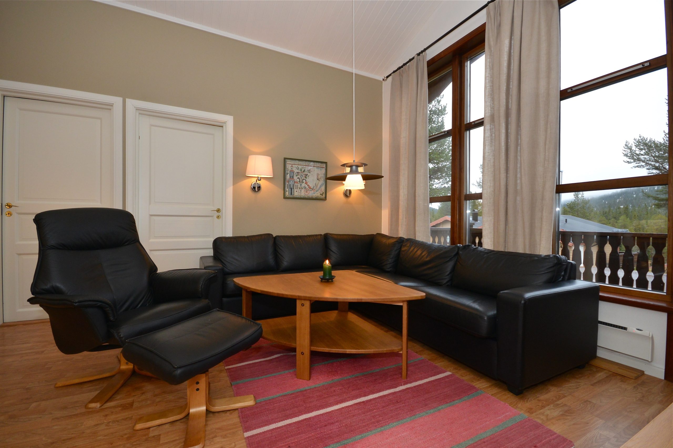 En hyggelig stue i Saalbach med en sektionssofa i sort læder og matchende lænestol, suppleret med et sofabord i træ og et rødstribet tæppe. Store vinduer med beige gardiner lukker naturligt lys ind, der minder om sommerdage under sommerferie Østrig. To lukkede døre pryder venstre væg.