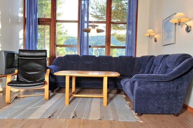 En hyggelig stue med en stor blå sektionssofa, en sort lænestol og et sofabord i træ på et gråt tæppe fremkalder charmen ved Wagrain. Store vinduer med blå gardiner lader naturligt lys strømme ind og viser træer udenfor. Væglamper tilføjer den indbydende stemning.