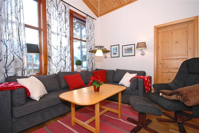 En hyggelig stue, der minder om en skiferie, med en grå sektionssofa prydet med røde og hvide hynder. Et træbord sidder i midten, toppet med et rødt blomsterarrangement. Værelset har store vinduer med træmønstrede gardiner og trævægge og -loft.