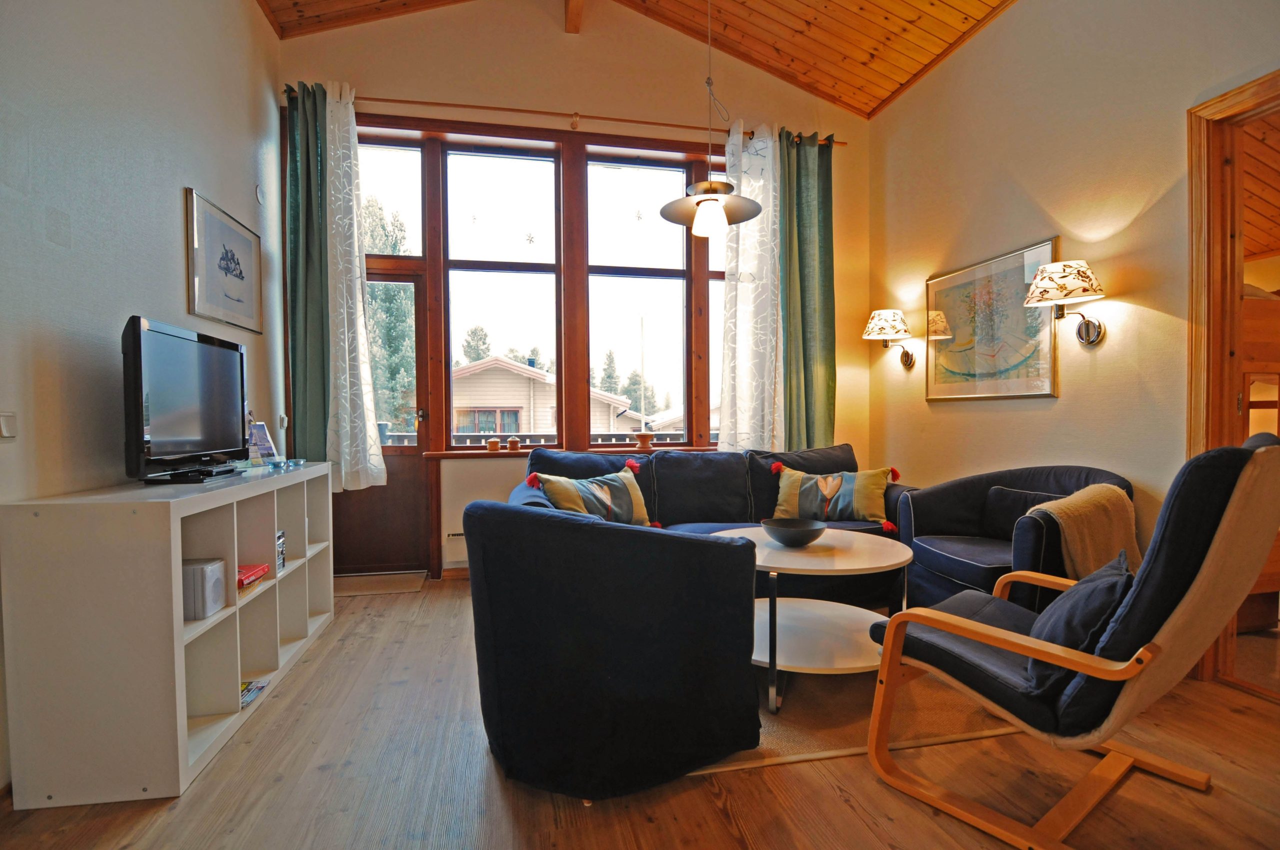Hyggelig stue med træloft, stort vindue, og trægulv. Inkluderer et tv på en hvid hylde, cirkulært sofabord, polstrede stole og en sofa. Væglamper og gardiner tilføjer varme. Med udsigter, der minder om Westendorfs charme under sommerferie Østrig, kan du næsten høre kaldet af skirejseeventyr.