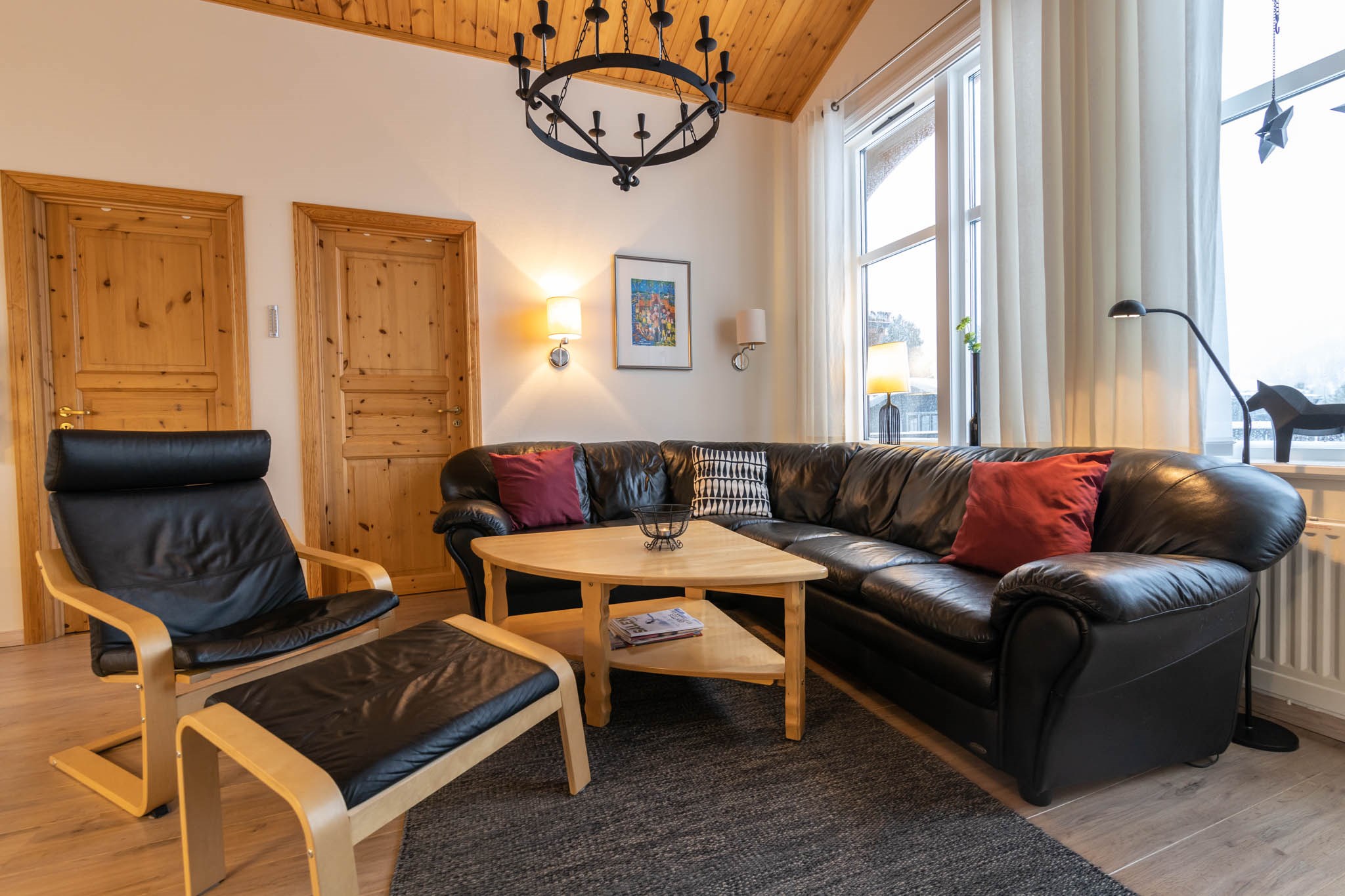 Hyggelig stue med sort lædersofa, røde hynder og tilhørende stol. Et sofabord i træ står på et gråt tæppe. Rummet minder om en Wagrain skiferie-hytte med trædøre, høje vinduer med lyse gardiner og en sort lysekrone hængende fra træloftet.