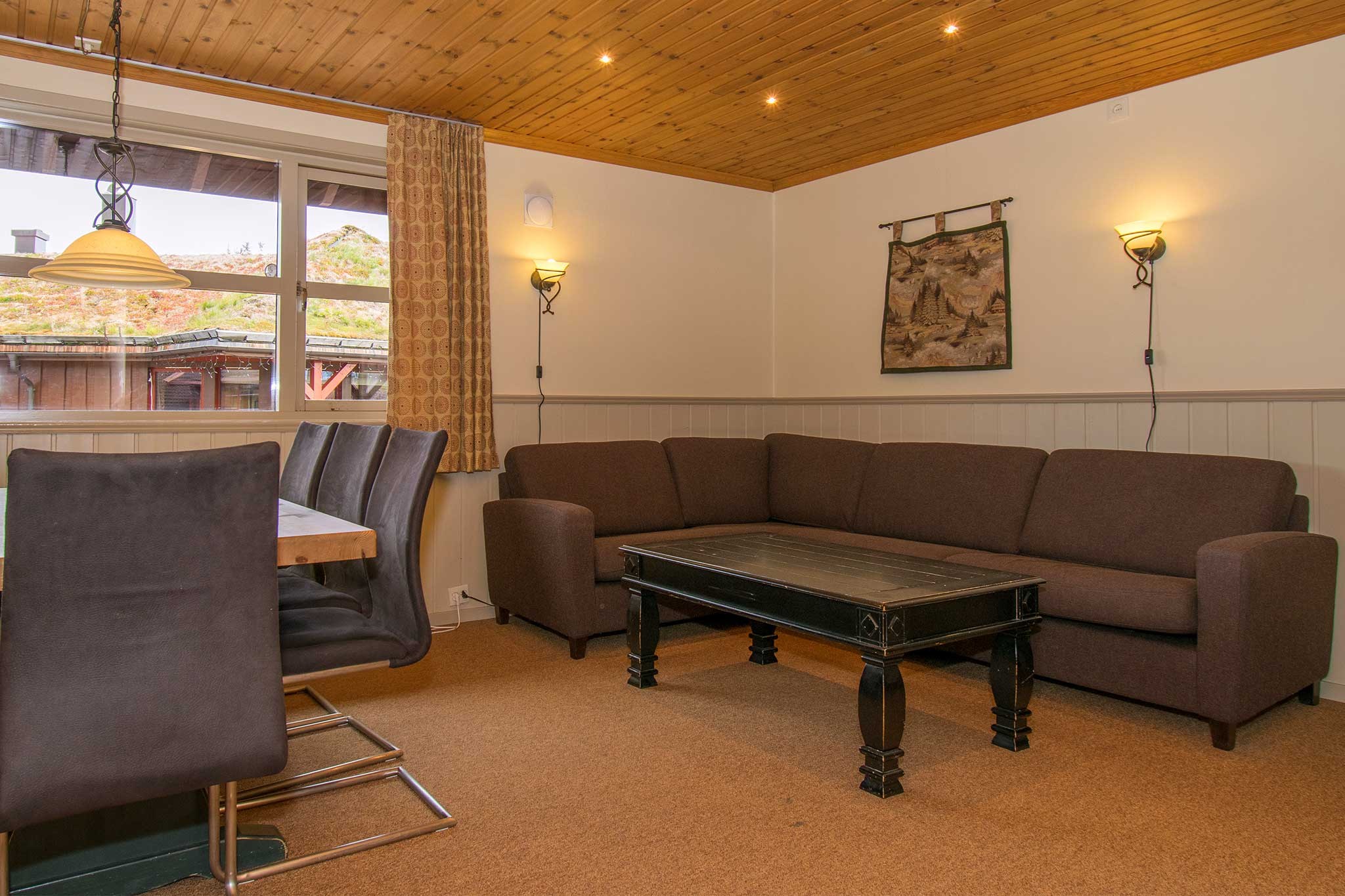 Hyggelig stue med brun sofa, sort sofabord og stole omkring et spisebord. Varm belysning fra væglamper og en loftslampe. Et stort vindue tilbyder en kahytsudsigt, der minder om et rustikt tilflugtssted i Wagrain. Vægkunst og loft med træpaneler fuldender den alpine charme.