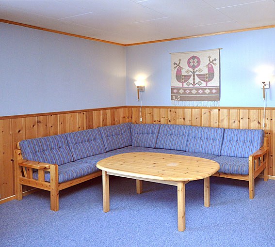 Et hyggeligt værelse med blåt gulvtæppe og vægge med træpaneler byder på en stor blå L-formet sofa og et rundt sofabord i træ, perfekt til at mindes om dine sommereventyr i Saalbach. To vægmonterede lamper oplyser et dekorativt tapet af vikingeskibe.