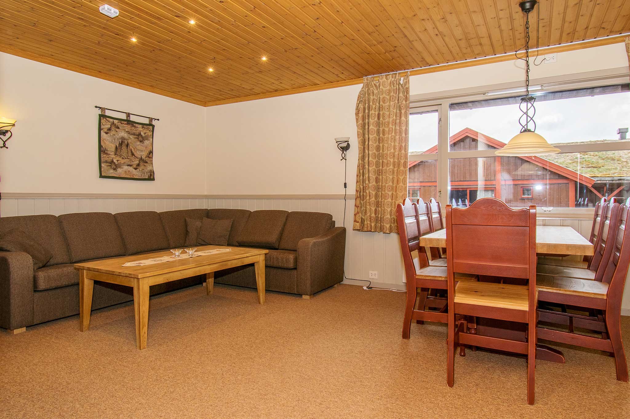 Et hyggeligt opholds- og spiseområde har en brun sektionssofa, sofabord i træ og spisebord med røde stole. Rummets træloft og beige vægge indrammer et vindue, der giver et glimt af en indbydende hytte, perfekt til at mindes om skiferieeventyr i Wagrain.