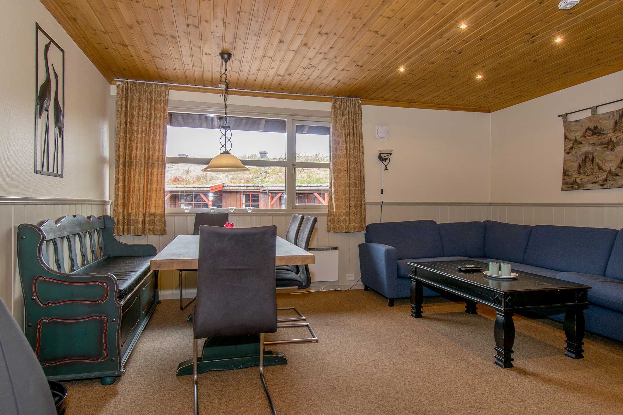En hyggelig stue med et træloft, der minder om Westendorf charme og et tæppebelagt gulv. Den har en blå L-formet sofa, et mørkt sofabord, en spiseplads med en bænk og stole, store vinduer med gardiner og dekorativ vægkunst.