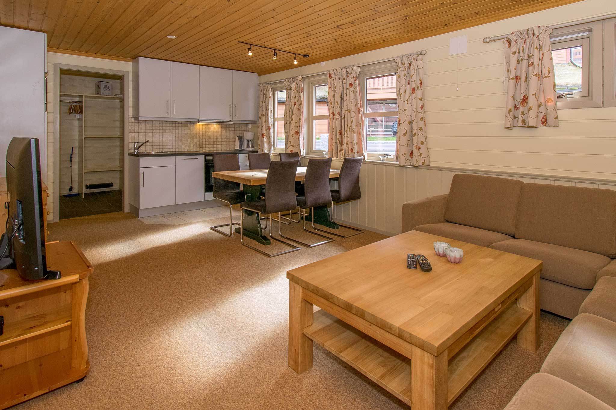 Hyggelig stue med træloft, der minder om en Wagrain skiferie. Lyse vægge omgiver den solbrune sofa og træsofabord med fjernbetjeninger. Et spisebord med sorte stole sidder i nærheden af køkkenområdet, med hvide skabe og en flisebelagt bagplade. Vinduer lukker naturligt lys ind.