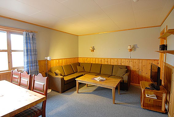 En hyggelig stue med træpaneler fremkalder et Saalbach-sommertilflugtssted med en stor brun sektionssofa, et sofabord i træ og et tv på en stativ. Et spisebord med stole er i forgrunden, mens et stort vindue med blå plaid gardiner tillader naturligt lys at strømme ind.