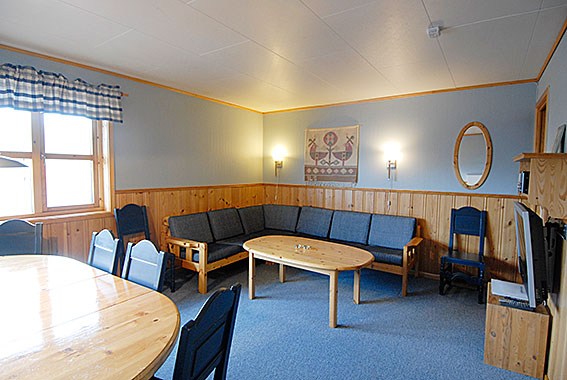 En hyggelig stue, der minder om et skiferiested i Westendorf, med træpaneler på vægge og blå gulvtæpper. Den har en stor grå sektionssofa, et sofabord i træ, vægmonterede lamper, et lille tv og et rundt spisebord med stole. Et vindue med et plaidgardin er til venstre.