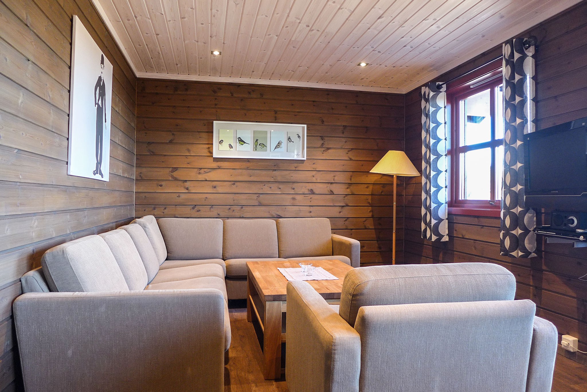 En hyggelig stue med trævægge inviterer til afslapning, med et sofasæt i beige stof, et sofabord i træ og en moderne gulvlampe. Kunstværker pryder væggene, og et vindue med mønstrede gardiner lyser rummet op. Et lille tv er monteret til højre, perfekt til afterski-fortællinger fra Westendorf eller Saalbach-eventyr.