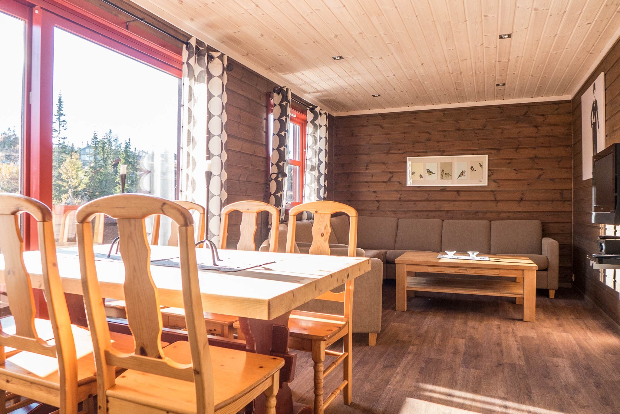 Et hyggeligt træhytteinteriør i Wagrain med spisebord og stole nær store rødrammede vinduer. En grå sofa står op ad væggen, og over den hænger et indrammet billede. Sollys strømmer ind, perfekt til en skiferiestemning.