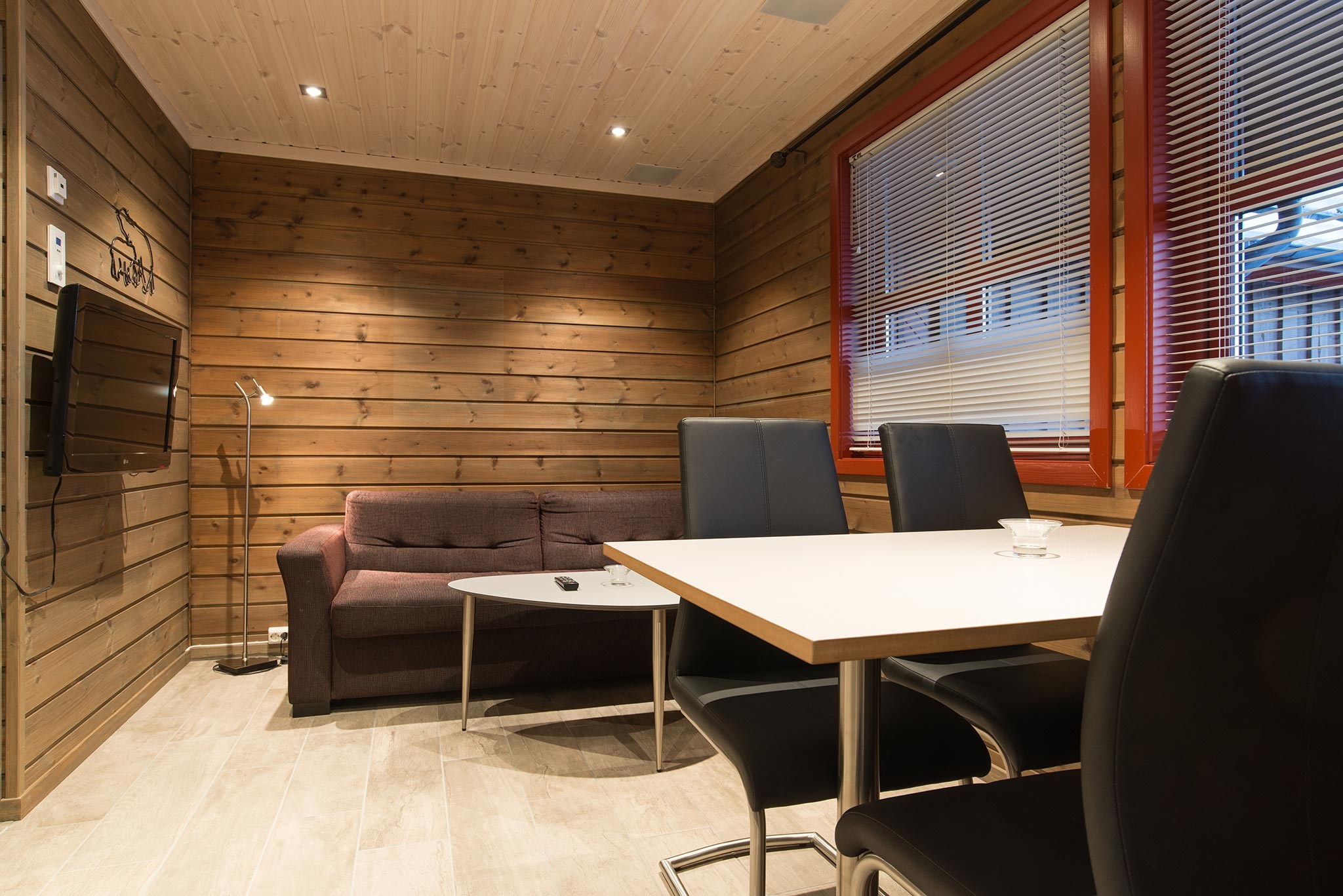 Hyggeligt træinteriør, der minder om charmerende retreater i Wagrain. Et lille opholdsområde har en brun sofa op ad væggen nær et tv. I forgrunden supplerer et moderne bord med sorte stole et stort vindue med persienner. Loftet med træpaneler tilføjer varme, perfekt til en sommerferie Østrig-stemning.