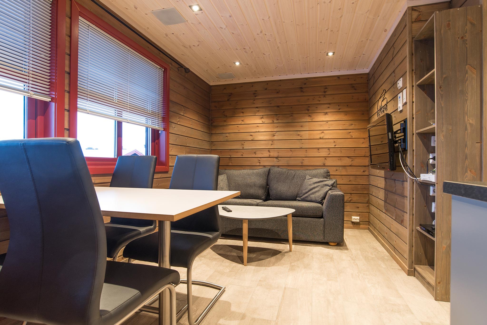 En hyggelig opholdsstue med trævægge og loft, der minder om en charmerende Saalbach skihytte. Den har en grå sofa, et lille sofabord og et spisebord med sorte stole. Rummet er veloplyst med indbygget belysning og naturligt lys fra to vinduer med persienner.