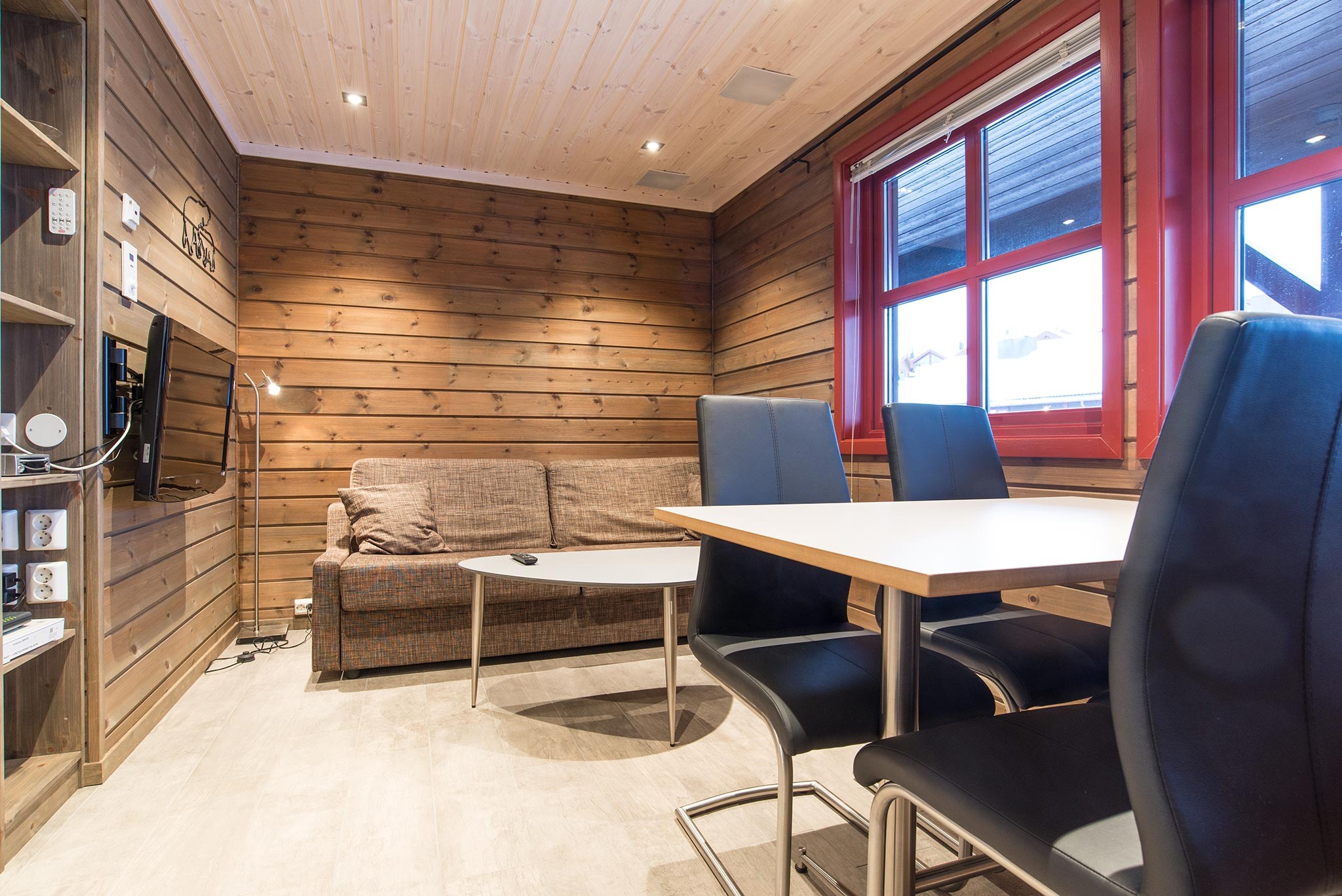 Hyggelig stue med trævægge og loft, der minder om en sommerferie i Westendorf. Et stort vindue med røde detaljer inviterer naturligt lys ind i sofaen, monteret tv, hvidt sofabord og spiseplads med bord og sorte stole. Perfekt til sommerferie Østrig-retreat!.