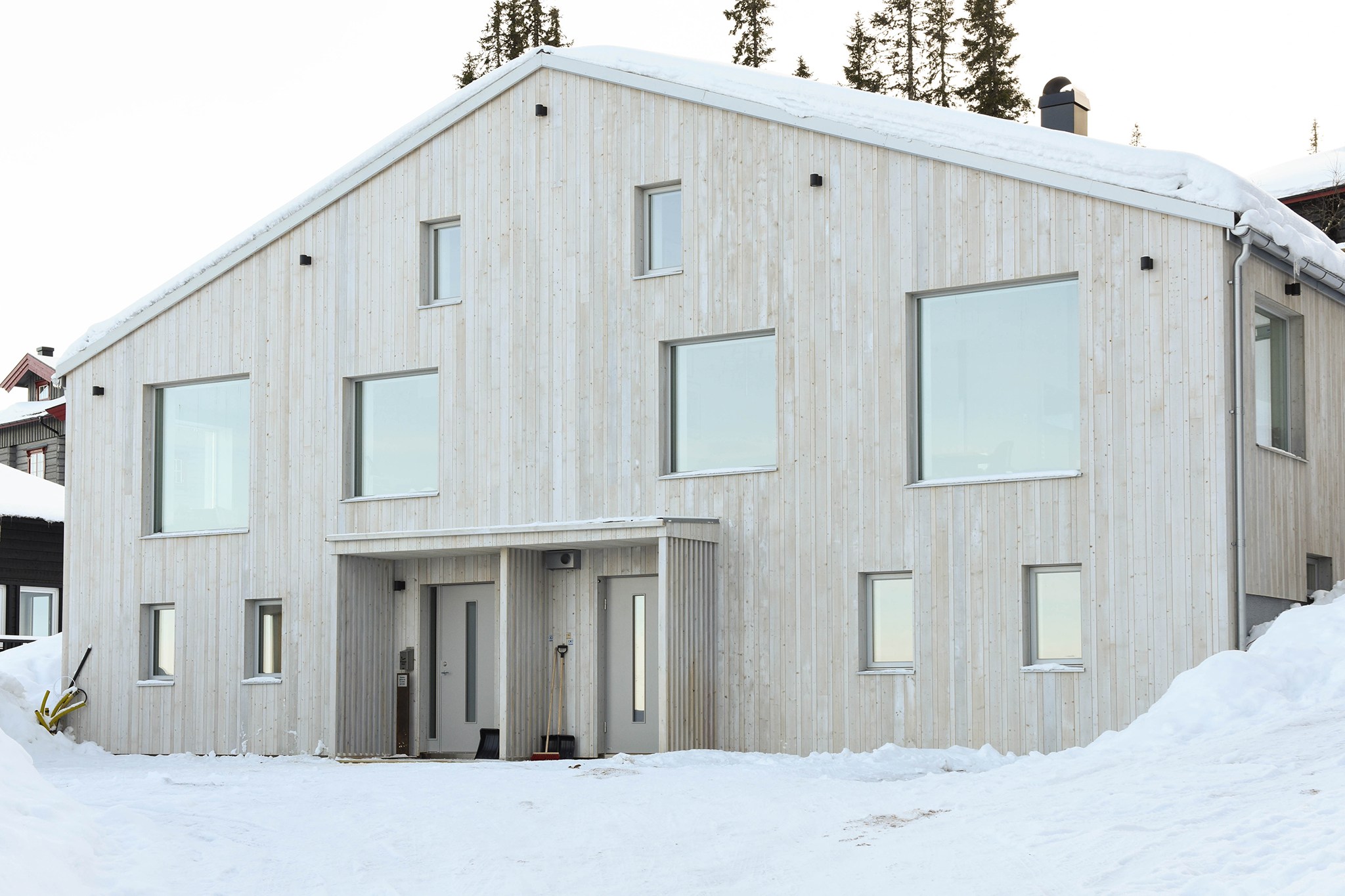 Et moderne lyst træhus med store vinduespartier er omgivet af sne, perfekt til en skiferie. Strukturen har et toppet tag og flere høje fyrretræer synlige i baggrunden, hvilket fremkalder en fredfyldt vinter indstilling, der minder om idylliske skirejser eventyr.