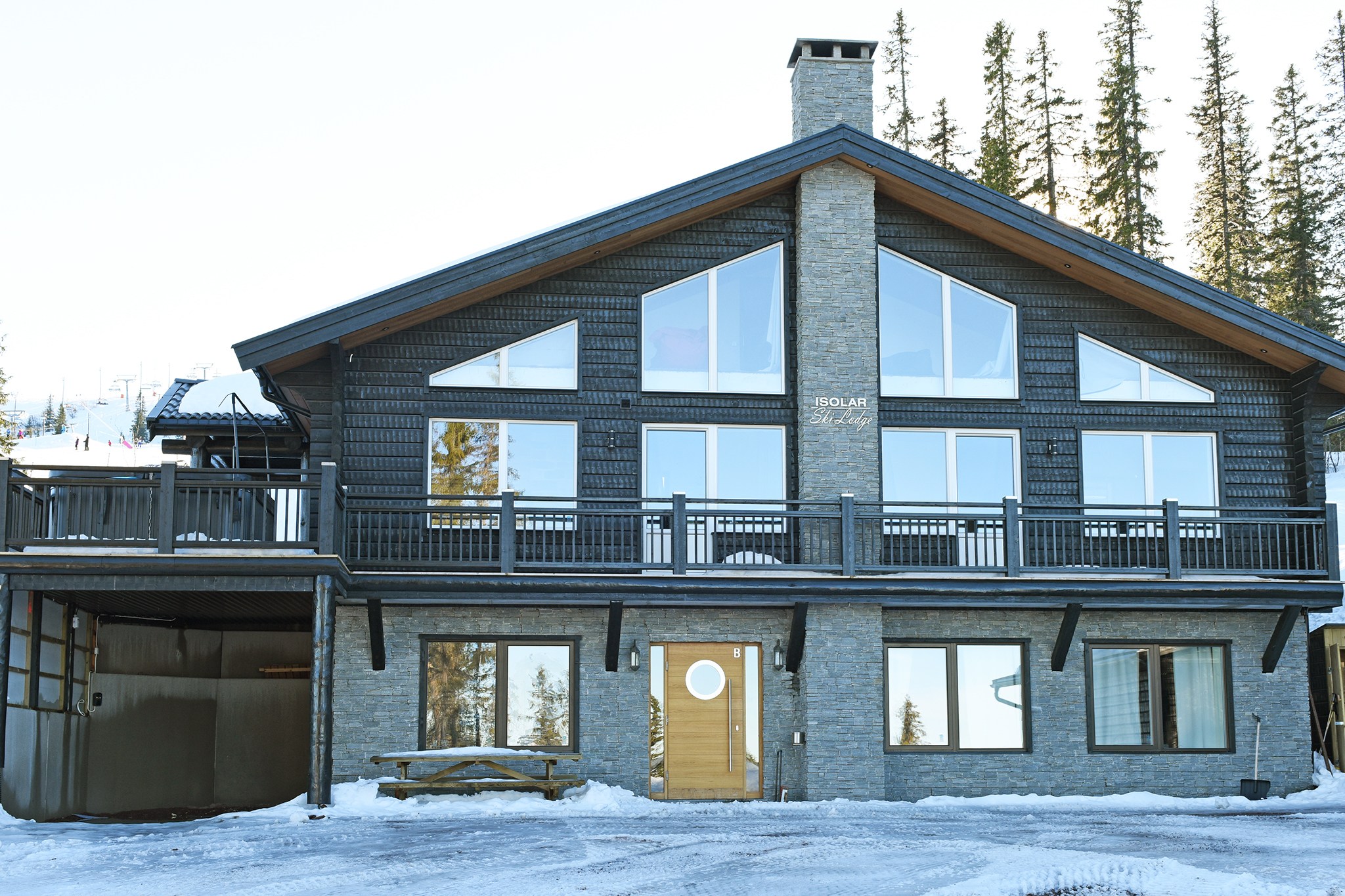 En stor, moderne hytte med en mørk træ- og stenfacade, med flere vinduer og en cirkulær trædør. Beliggende i det sneklædte landskab i Wagrain, Østrig, tilbyder det en fantastisk udsigt med høje stedsegrønne træer i baggrunden, perfekt til en unik sommerferie Østrig-oplevelse.