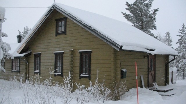 Et snedækket træhus med et stejlt, skråt tag i Saalbach udstråler et hyggeligt udtryk. Omgivet af snefyldte træer og buske, står det nær en rød pæl under en overskyet himmel, perfekt til en skiferie flugt.