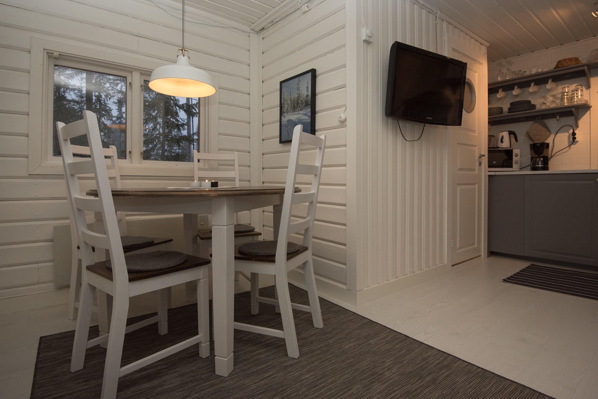 En hyggelig spiseplads i en træhytte, perfekt til en skiferie flugt. Med hvide vægge og loft har den et rundt bord med fire stole under en pendel og et tv monteret på væggen. I baggrunden er der et tekøkken med hylder og hvidevarer - et lille stykke Wagrain-charme.