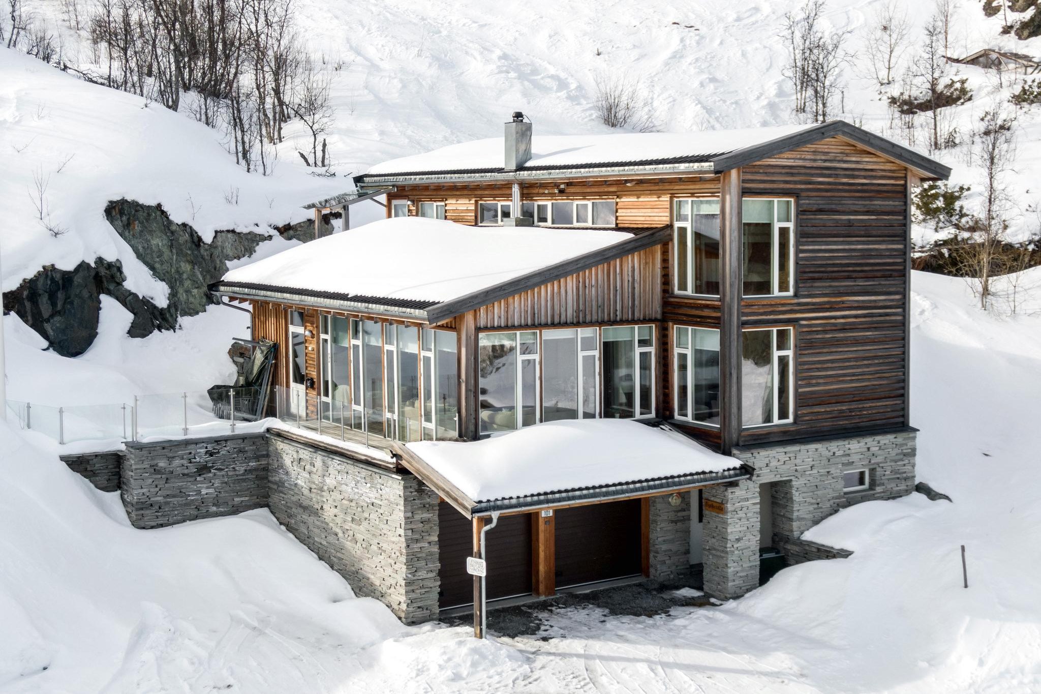 En moderne to-etagers hytte med store vinduer, træ og sten udvendigt er beliggende i det snedækkede landskab i Saalbach. Huset har et skrå tag, og den lyse sne står i kontrast til træets varme toner – perfekt til en hyggelig skiferieferie.