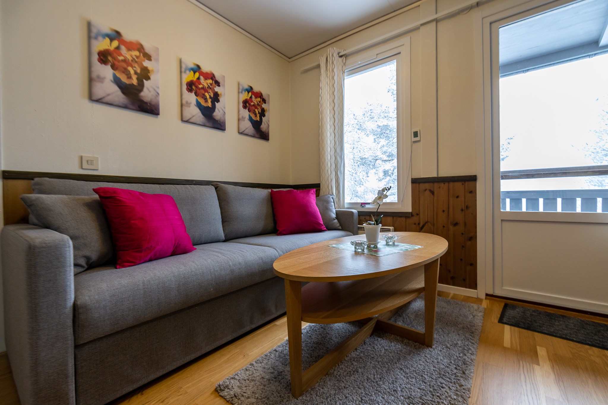 En hyggelig stue med en grå sofa prydet med to knaldrøde puder, der minder om dit skiferieophold i Wagrain. Tre blomstermalerier hænger på væggen. Et sofabord i træ hviler på et gråt tæppe ved vinduet og lukker lys ind, der lysner rummet op.
