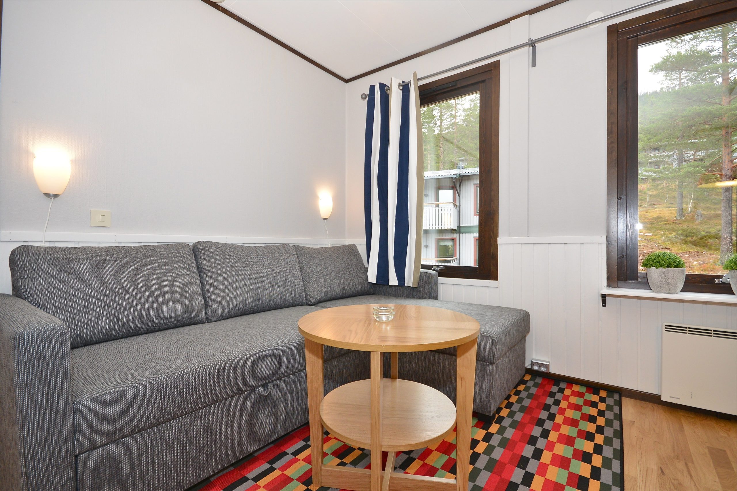 En hyggelig stue med en grå sofa, et lille rundt bord i træ og store vinduer med udsigt, der minder om det fredfyldte grønne område i Saalbach. Gulvet kan prale af et farverigt mønstret tæppe, og en lille plante pryder vindueskarmen ved siden af stribede gardiner.