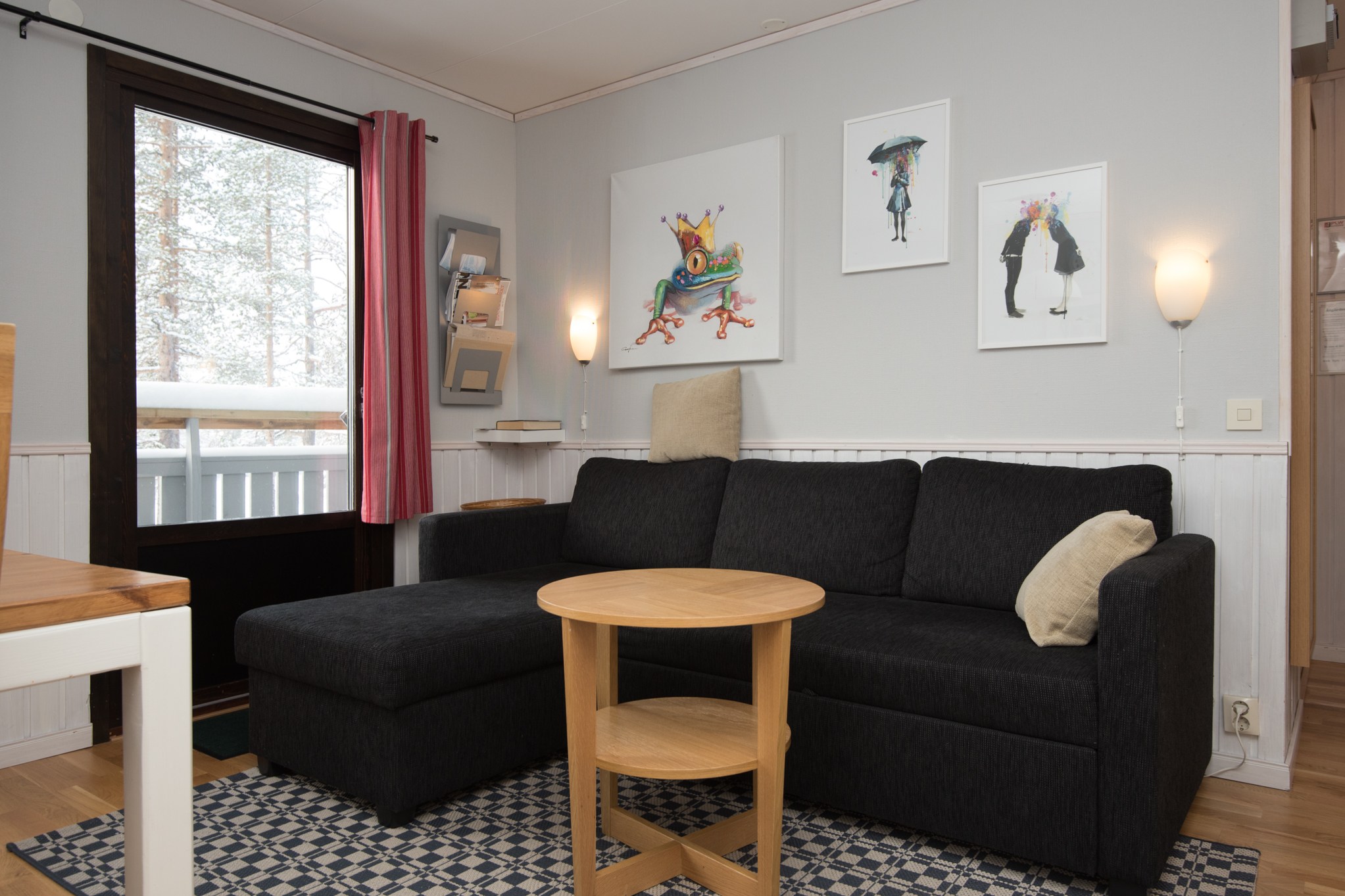 En hyggelig stue har en sort sektionssofa med en beige pude, et rundt sofabord i træ og eklektisk kunst, der minder om Saalbachs charme. Et lille spisebord i træ er delvist synligt, og en dør åbner udad. Et blødt tæppe dækker gulvet.