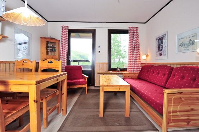En hyggelig stue, der minder om en hyggelig hytte, har et spisebord i træ med stole, en rød polstret sofa og en matchende lænestol. Et sofabord af træ står på et tæppe i midten, mens lyset filtrerer gennem rødternede gardiner og fremkalder varmen fra afterski-aftener i Wagrain.