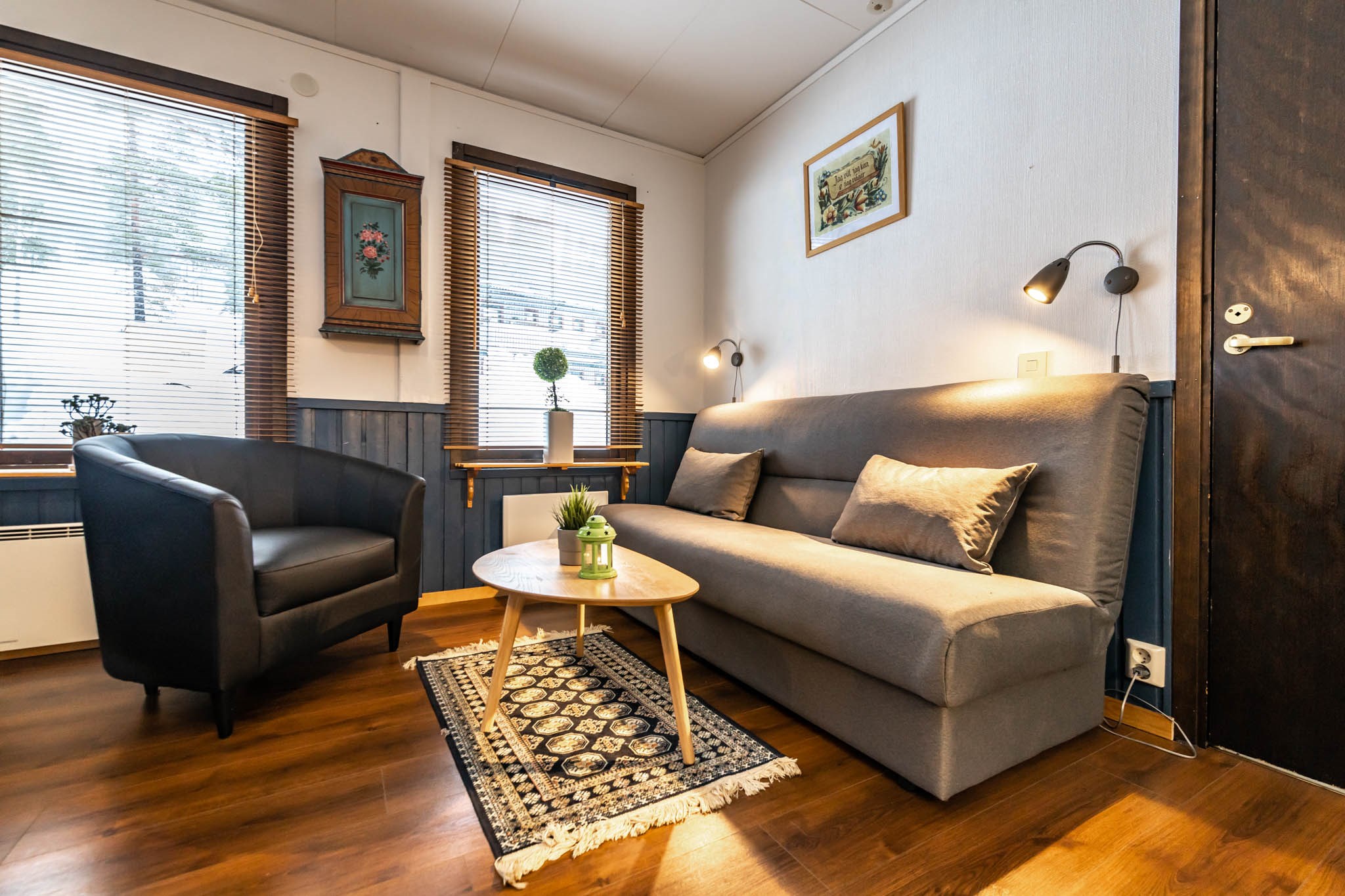 En hyggelig stue, der minder om et Wagrain-retræte, med en grå sofa, sort lænestol og et ovalt sofabord i træ på et mønstret tæppe. To små lamper over sofaen oplyser rummet. Store vinduer med persienner og vægdekorationer fuldender denne charmerende flugt.
