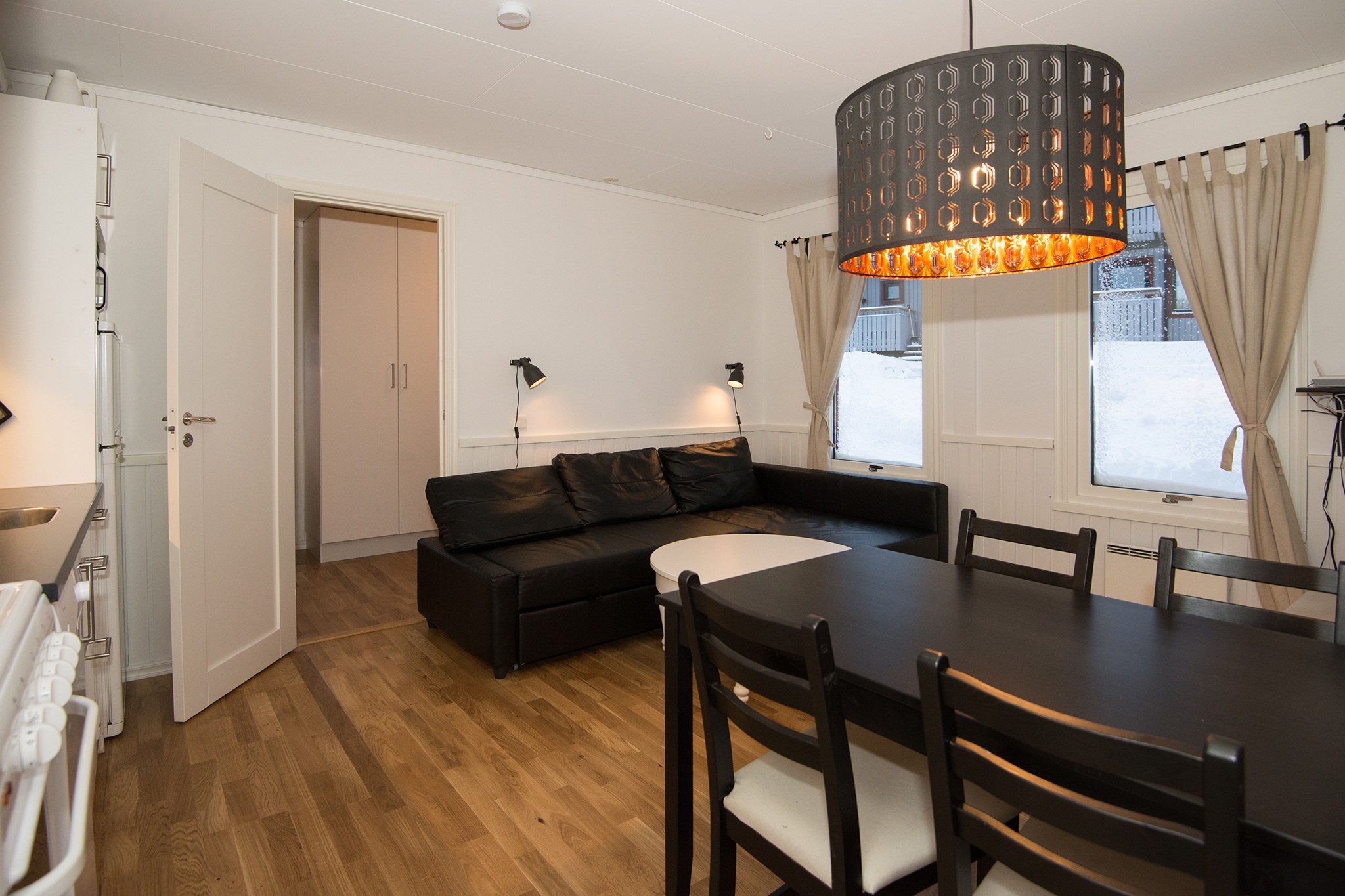 En hyggelig stue i Westendorf med sort hjørnesofa, rundt hvidt sofabord og sort spisebord med stole. En moderne pendel hænger over trægulve. Gardiner pryder vinduerne med en dør, der fører til et andet rum, der minder om en perfekt skiferie-ferie.