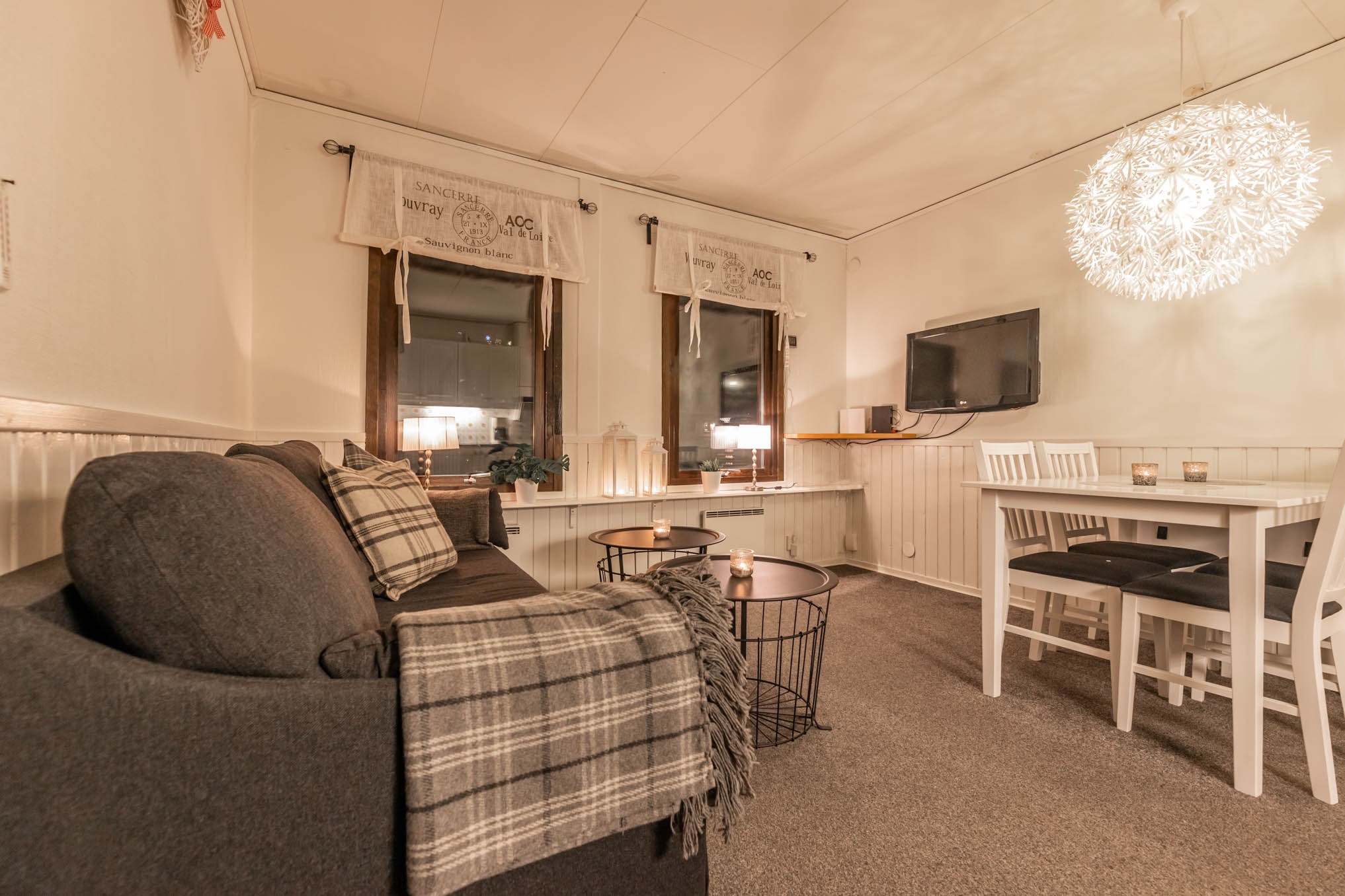 Hyggelig stue med en grå sofa prydet med plaid tæpper. Et moderne hvidt spisebord med sorte stole er i nærheden. Dæmpet belysning fra en dekorativ pendel skaber en varm atmosfære, der minder om aftenen i Westendorf. To store vinduer har trykte gardiner.