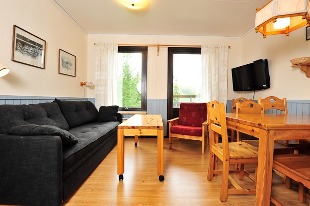 En hyggelig stue, der minder om en Wagrain-ferie, med en sort sofa, rød lænestol og sofabord i træ. Til højre er en spiseplads klar til en sommerflugt i Østrig. Et tv pryder væggen, mens store vinduer med gardiner bader trægulvet og kunstudsmykkede vægge i naturligt lys.