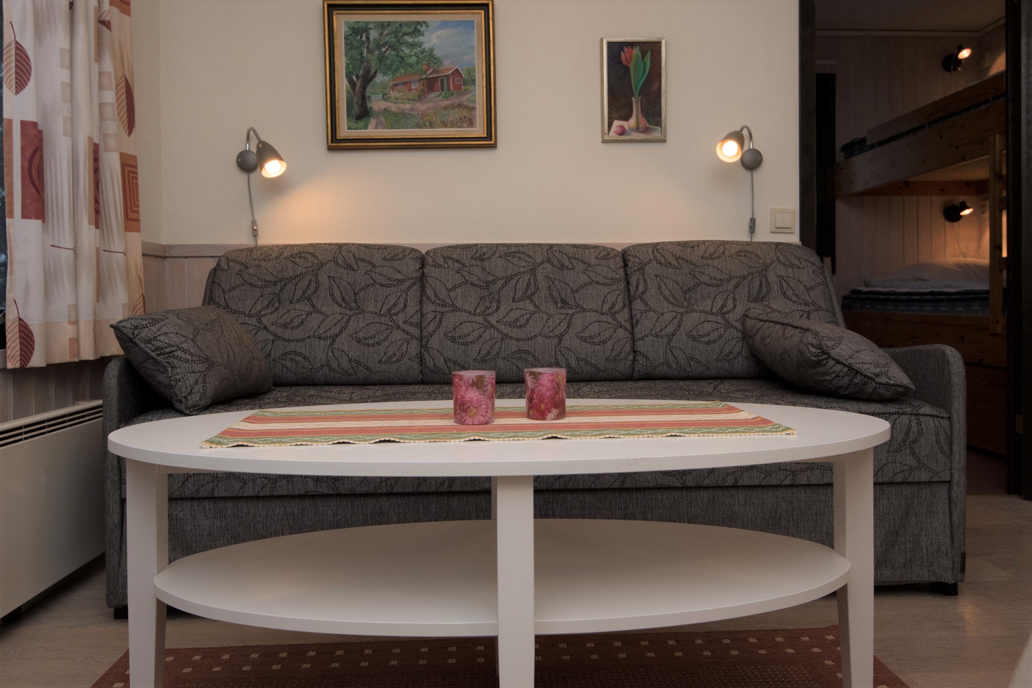 En hyggelig stue i et Wagrain-retræte har en mønstret grå sofa mod væggen, prydet med to malerier. Et hvidt ovalt sofabord holder lyserøde stearinlys på en farverig løber. Blød belysning fra væglamper giver varme, mens køjesenge antyder overnatning perfekt til skirejser eller sommerferie i Østrig.