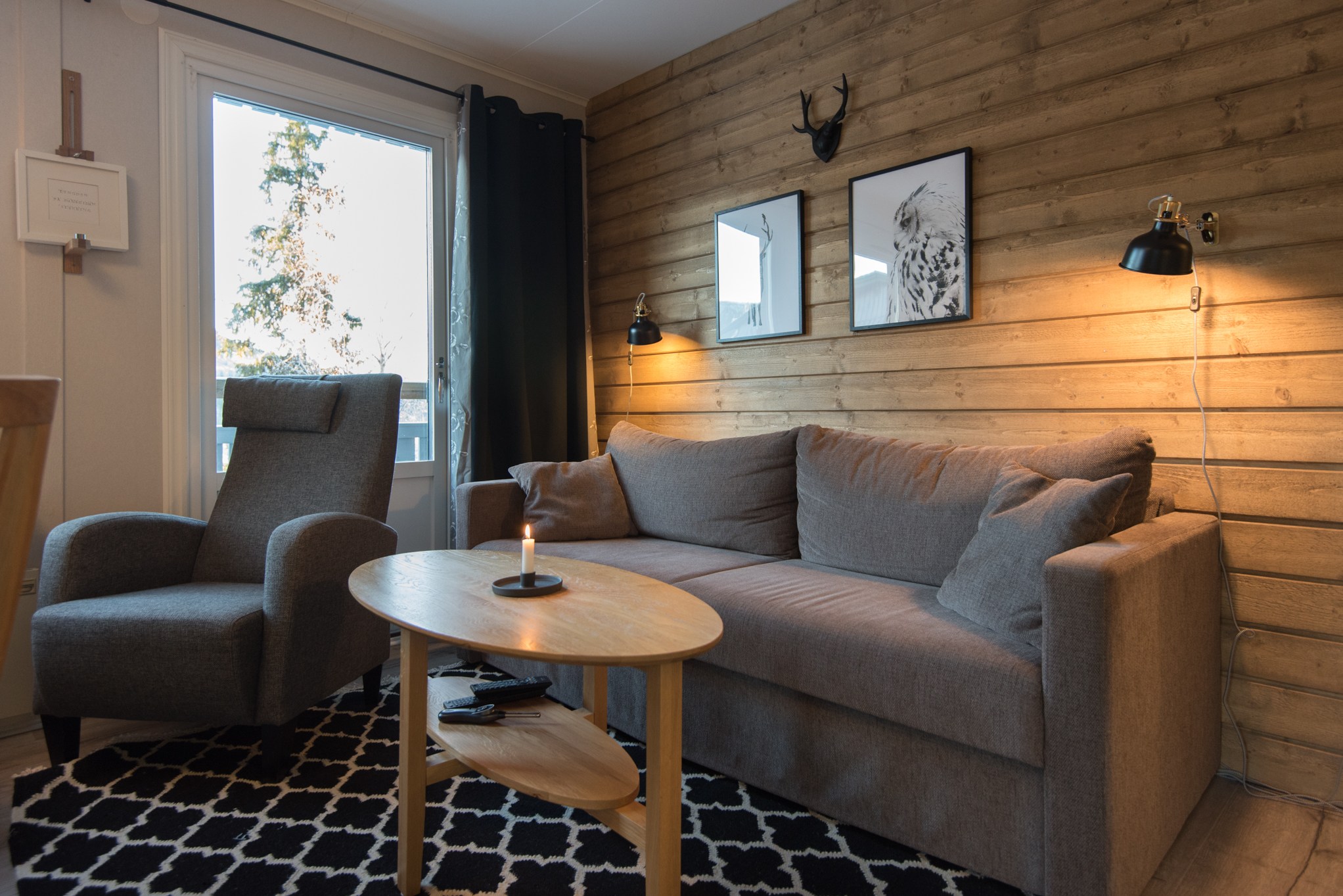 En hyggelig stue med en træ-accentvæg har en grå sofa og lænestol, der minder om en Westendorf-hytte. Et lille sofabord i træ med et stearinlys og sort-hvide indrammede kunstværker tilføjer charme, mens varm belysning forstærker den indbydende stemning, perfekt efter en dag på din skiferie.