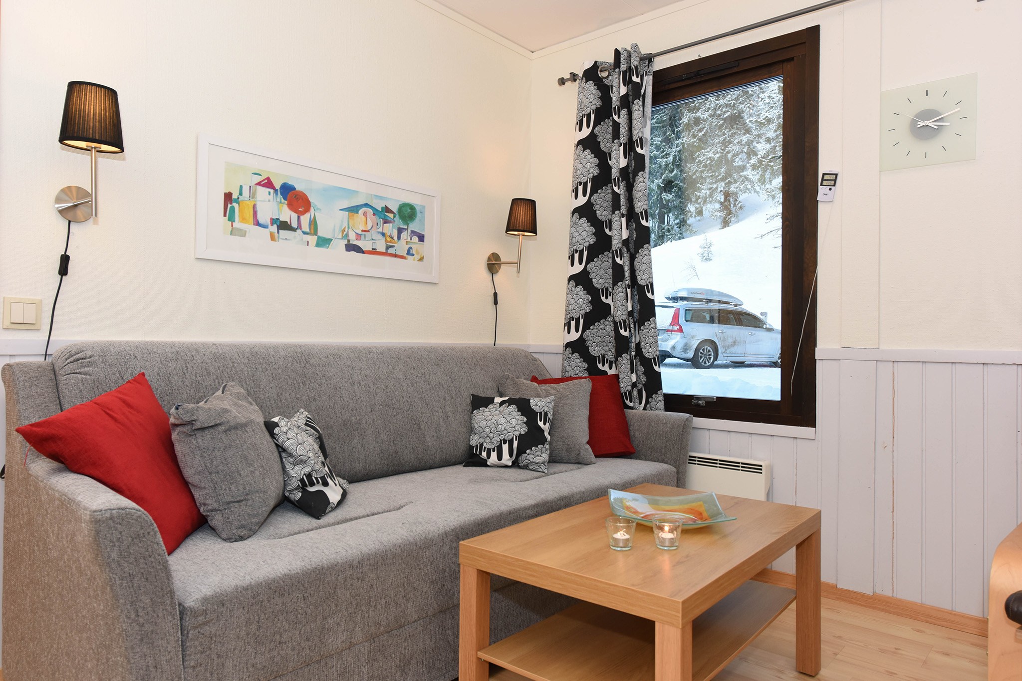 En hyggelig stue med en grå sofa prydet med røde og grå puder med fåredesign. Et sofabord af træ rummer en bog og stearinlys. Et farverigt maleri, der minder om Westendorfs fantastiske udsigter, hænger over sofaen, mens et vindue afslører et snelandskab udenfor.