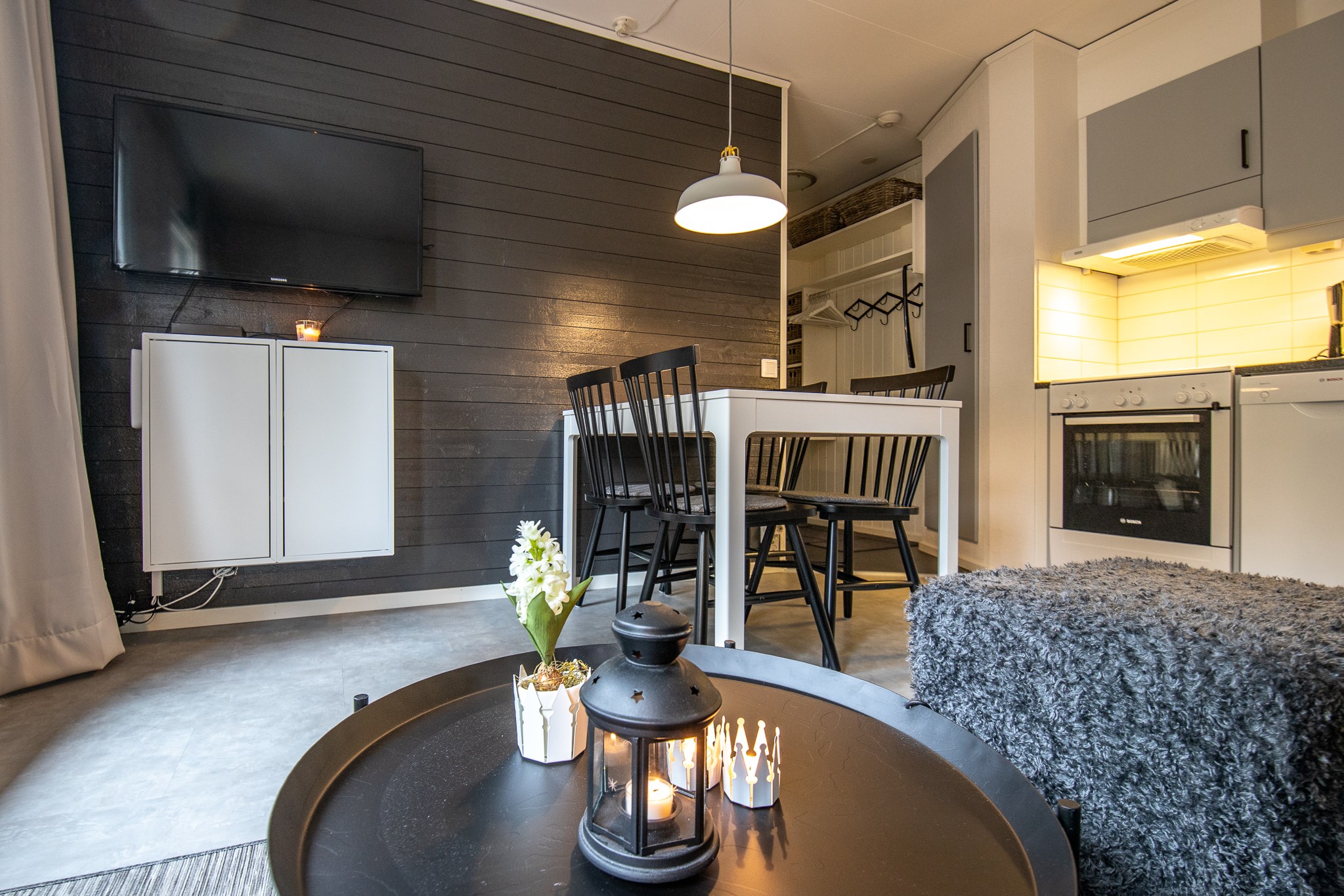 Et moderne opholdsområde med en sort accentvæg med et monteret tv fremkalder det slanke design af populære skihytter i Saalbach. I nærheden supplerer et spisebord med sorte stole køkkenets hvide skabe. Et sort sofabord prydet med en lanterne og stearinlys tilføjer hyggelig charme.