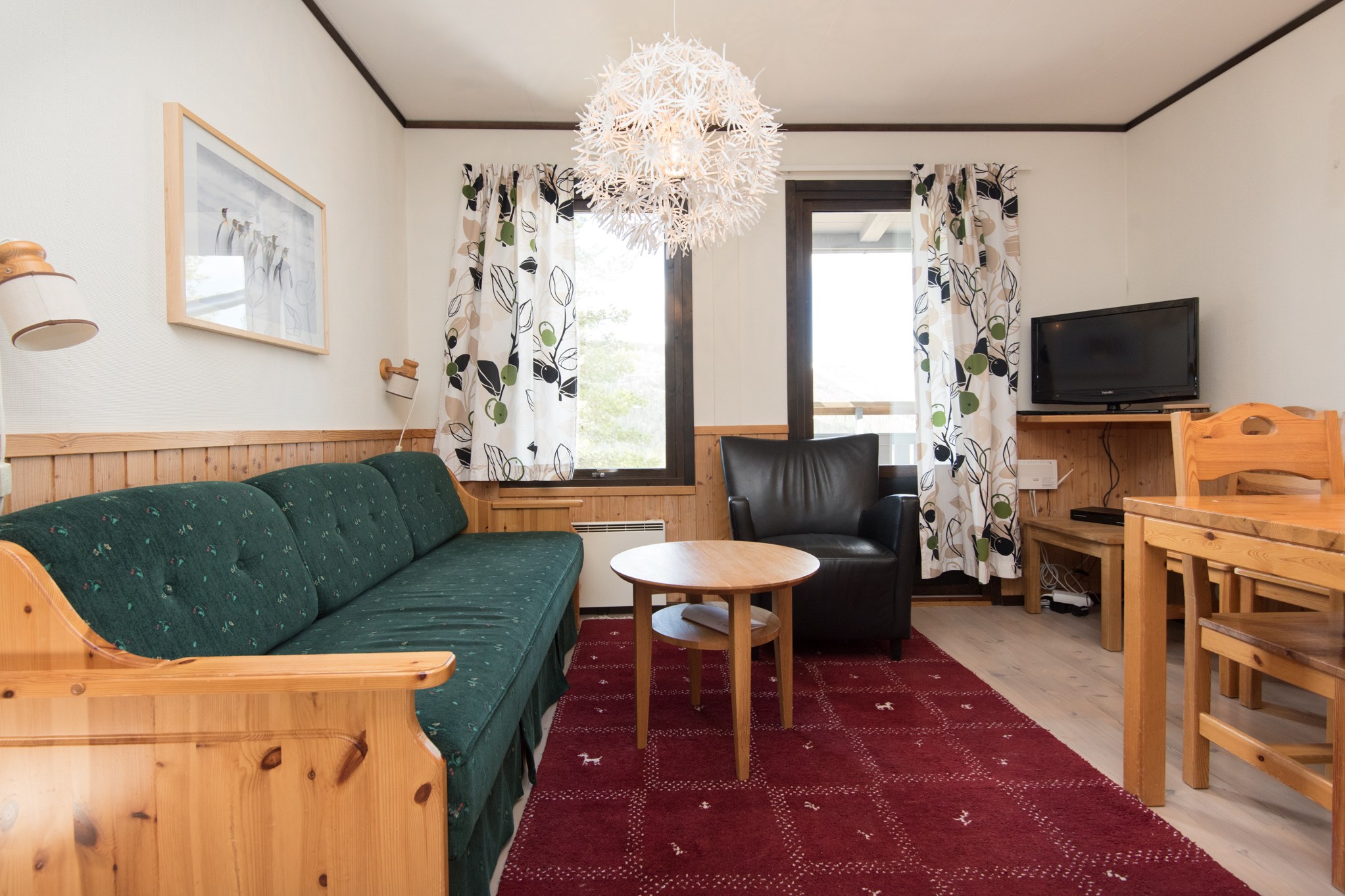 En hyggelig stue med træmøbler, herunder en mørkegrøn sofa, sort lænestol, lille rundt bord, og et træ spisebord med stole. En unik lysekrone og blomstergardiner fanger charmen ved en Westendorf skiferie. Et fjernsyn er placeret på et træskab til afslappende aftener.