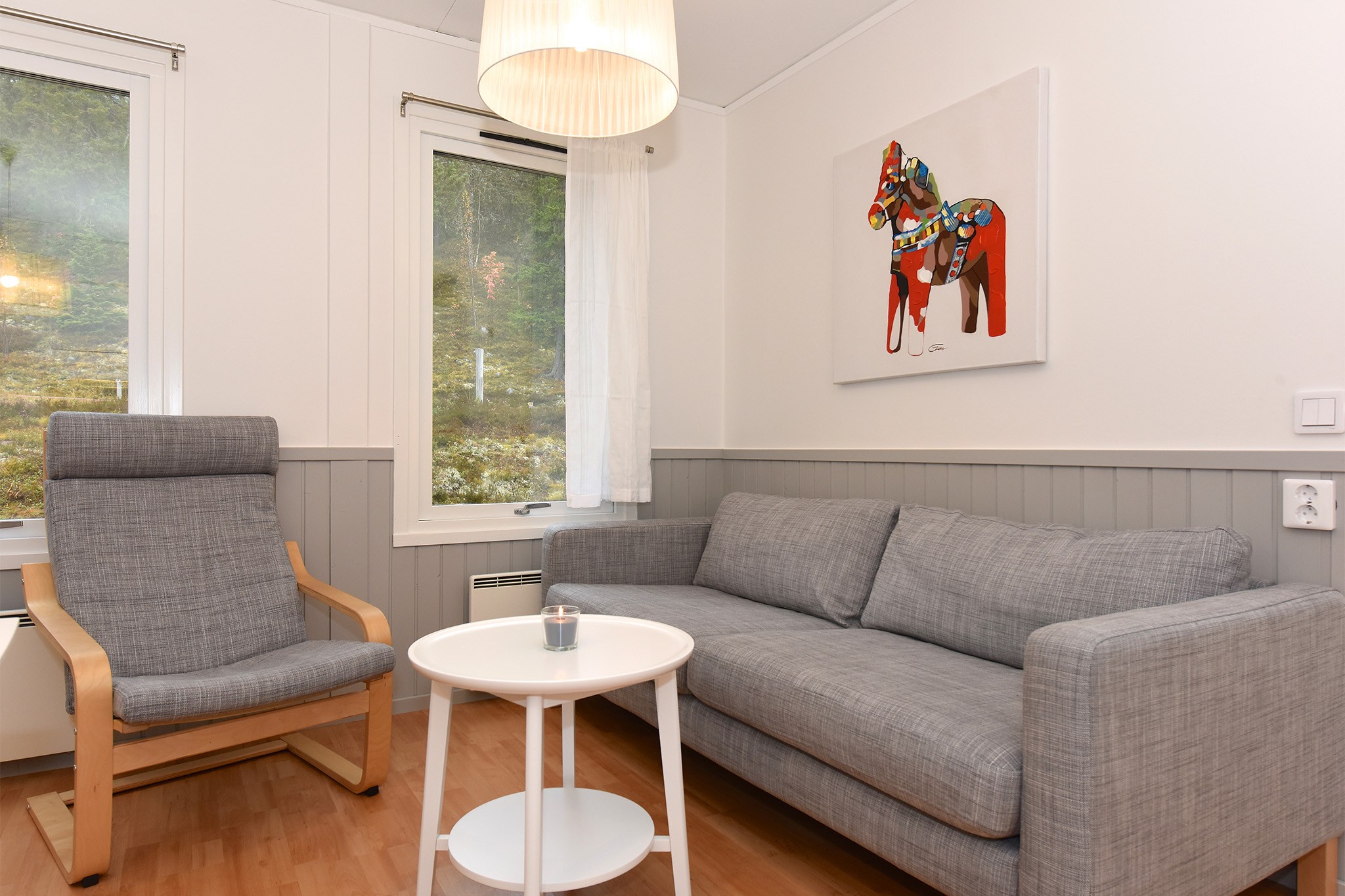 En moderne stue med en grå sofa og matchende lænestol, et rundt hvidt sidebord med et lille lys og et maleri af en farverig hest. Vinduerne byder på udsigter, der minder om de fredfyldte landskaber i Westendorf.