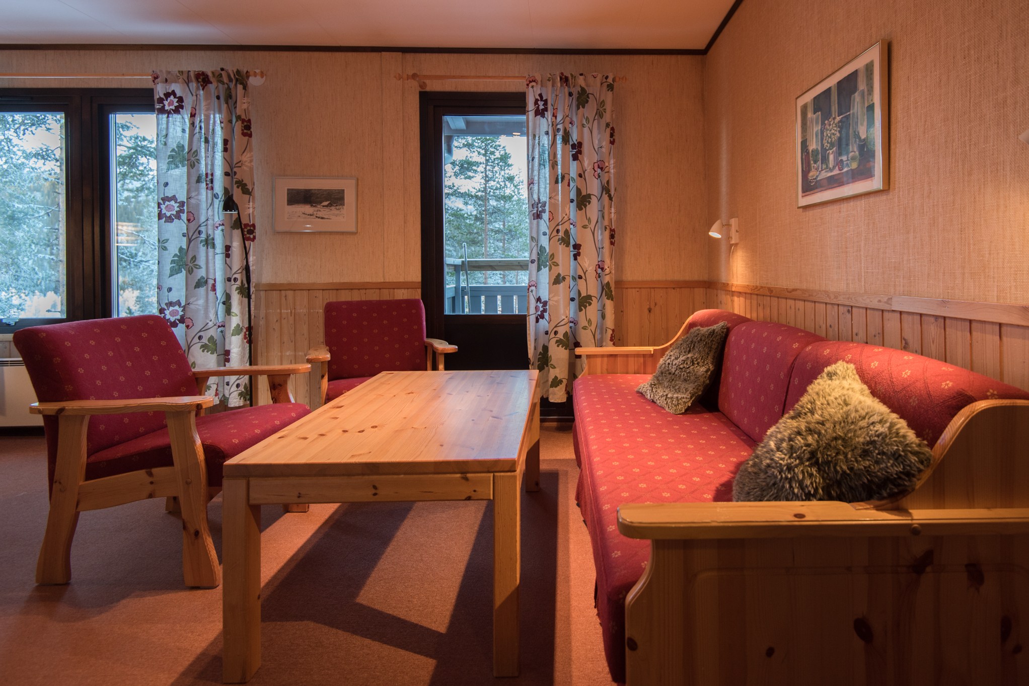 En hyggelig stue med varm belysning, med et sofabord i træ og en rød polstret sofa med hynder - en perfekt flugt, der minder om en Saalbach-skirejser. Blomstergardiner indrammer vinduet og døren, mens væggene er prydet med indrammet kunst.