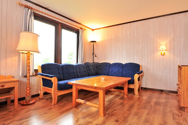 En hyggelig stue i Westendorf med trævægge og trægulve, med en blå polstret sofa, et sofabord i træ, en gulvlampe og en lille vægmonteret lampe. To store vinduer med gardiner tillader naturligt lys at oplyse denne perfekte flugt efter en dag på pisterne.
