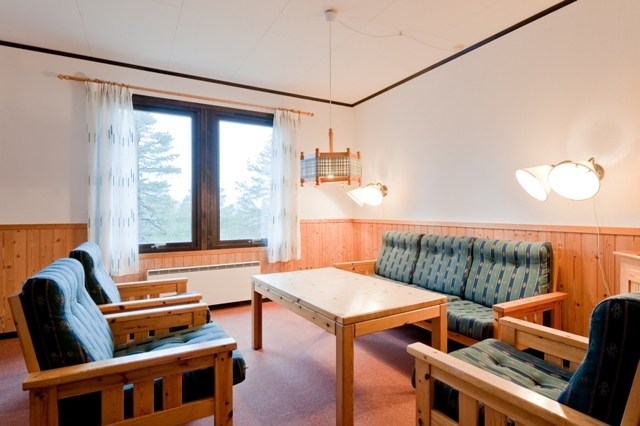 Et hyggeligt værelse perfekt til din sommerferie Østrig, med et træbord med tilhørende stole, en behagelig sofa og to væghængte lamper. Lyse træpaneler fremhæver væggene, mens et stort vindue med rene gardiner afslører den fredfyldte skønhed af Wagrains træer udenfor. En loftslampe lyser over bordet.