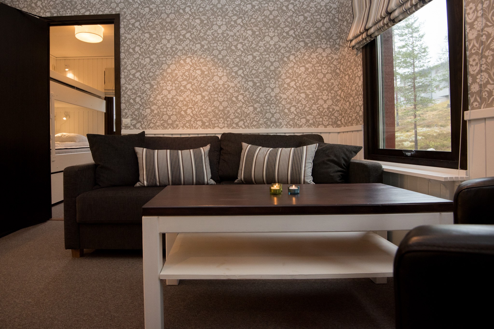 En hyggelig stue med en mørkegrå sofa prydet med stribede puder inviterer dig til at slappe af, der minder om en sommer i Westendorf. Et sofabord af træ står foran, og et vindue viser træer udenfor. Det blomstermønstrede tapet tilføjer charme, med et glimt af et soveværelse gennem døråbningen.