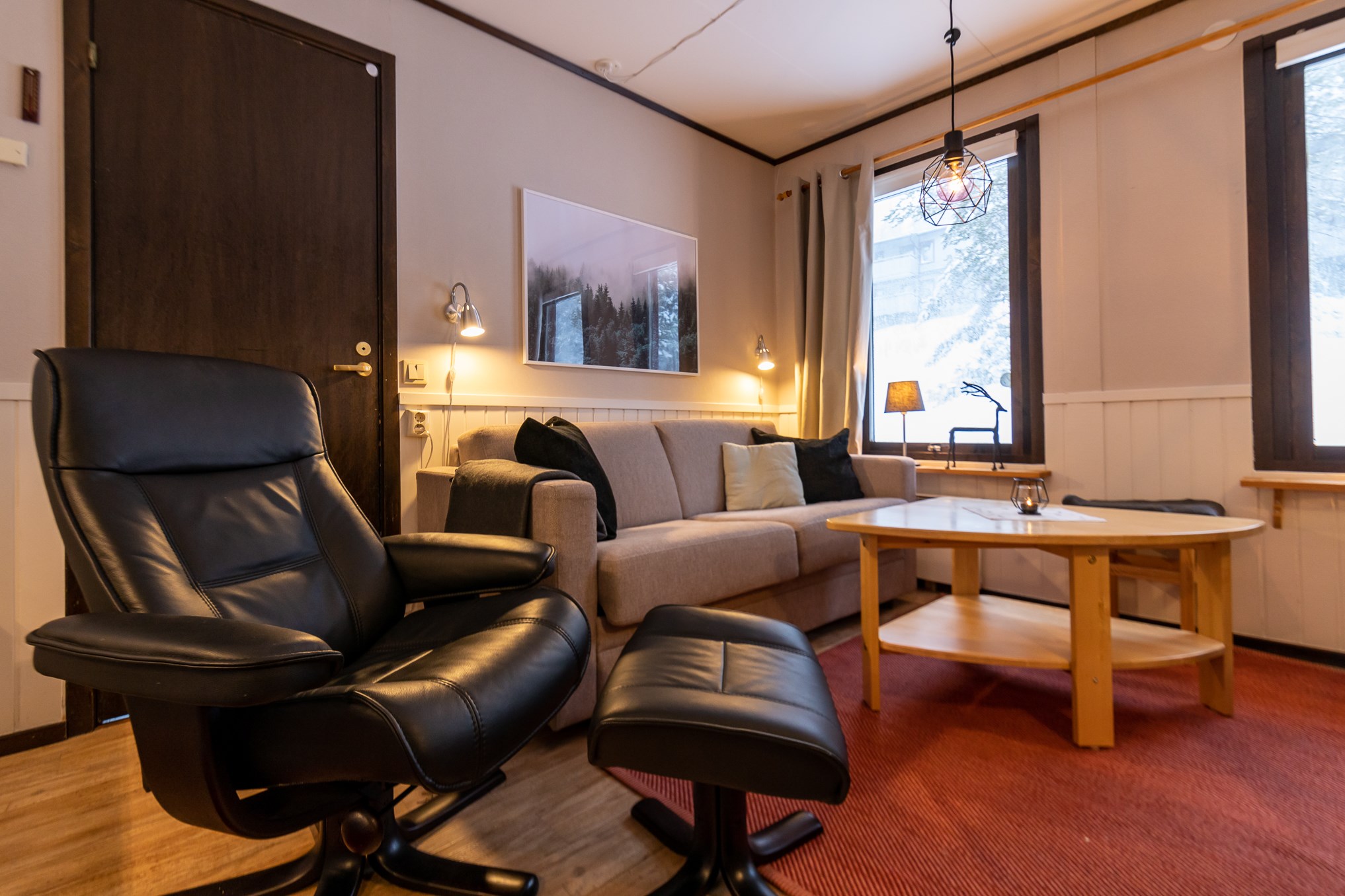 En hyggelig stue, der minder om et charmerende skiferiested med en beige sofa og hynder, sort læderstol med skammel og et sofabord i træ. Den varme belysning fra en hængelampe ligner det alpine skær i Wagrain, der kaster bløde skygger på indrammet kunstværk ved vinduesvæggen.