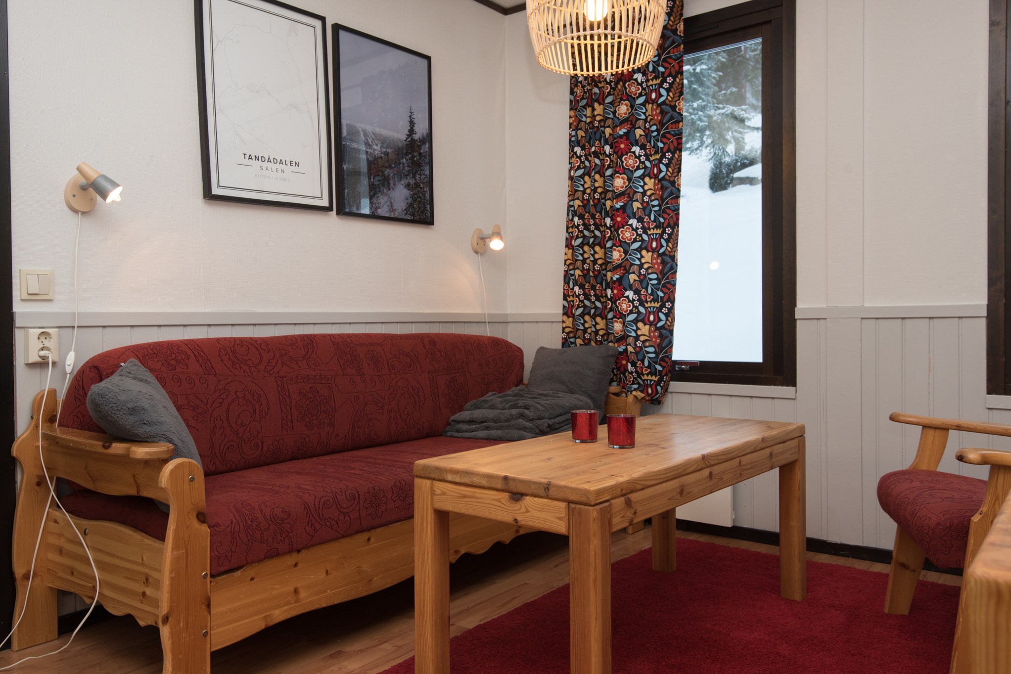 Hyggelig stue med træmøbler, inspireret af charmerende Wagrain. Rummet har en rød sofa og lænestol, mens et sofabord i træ rummer røde stearinlys. Vægkunst og et farverigt gardin tilføjer flair, og ovenover hænger en vævet pendel, der giver et varmt skær, der minder om din sommerferie Østrig.