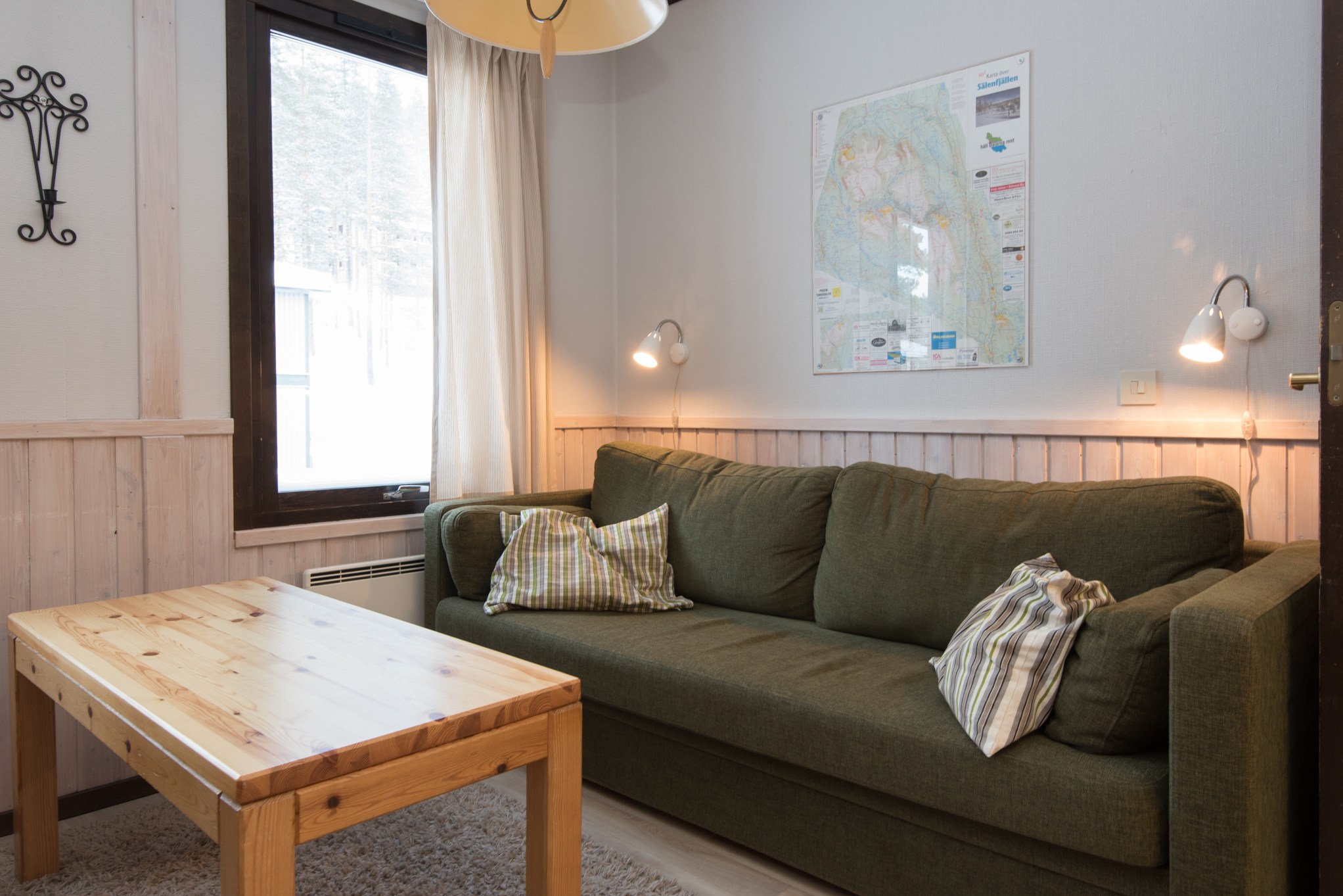 En hyggelig stue med en grøn sofa, plaidpuder og et sofabord i træ. To små væglamper giver varmt lys, der minder om at slappe af efter en dag i Saalbach. Et stort vindue med et gardin er til venstre, og et kort er vist på væggen over sofaen.